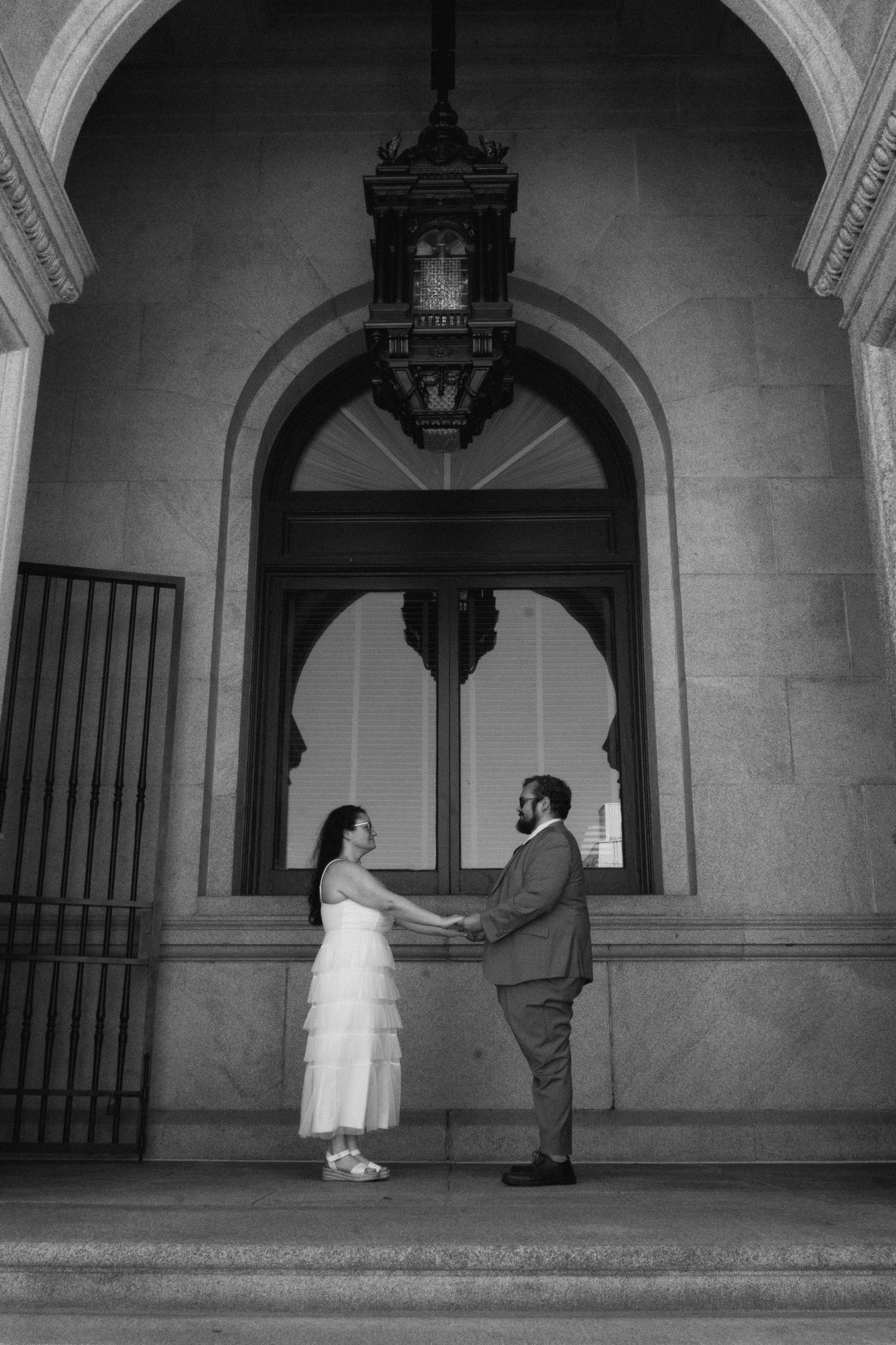 Black and white photo of a couple holding hands in wedding attire beneath an ornate arch with a vintage lantern.
