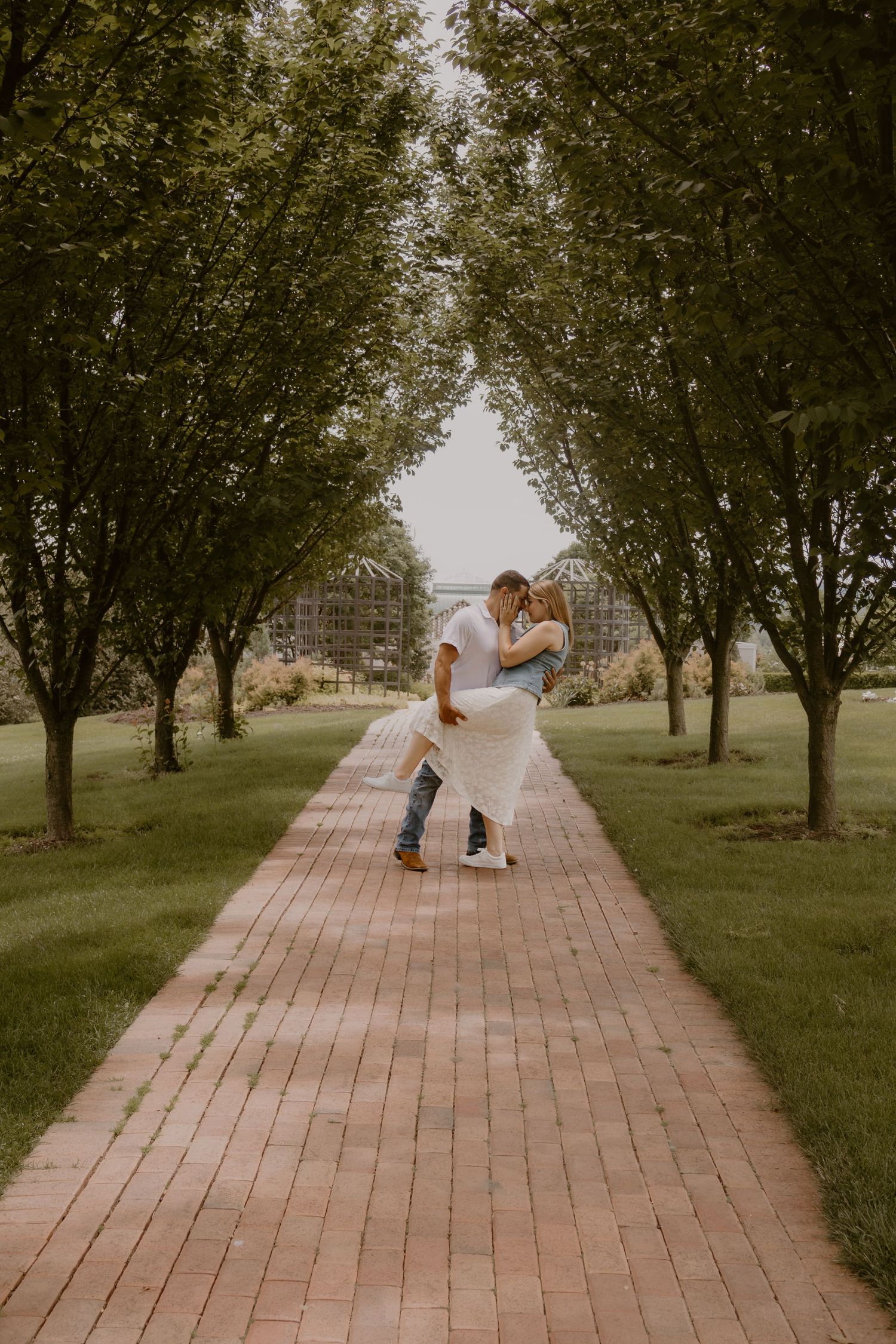 Couple sharing a romantic dip kiss on a brick pathway lined with lush green trees in a park setting.