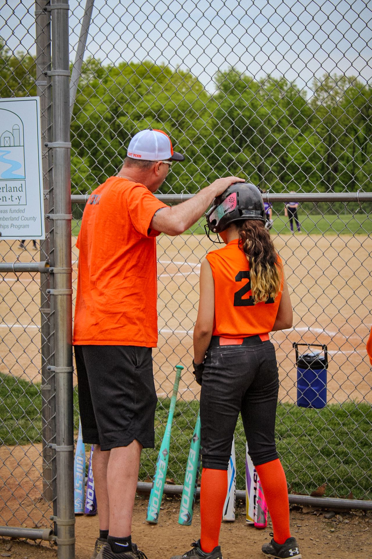 A coach in orange adjusts equipment for a softball player in matching orange uniform.