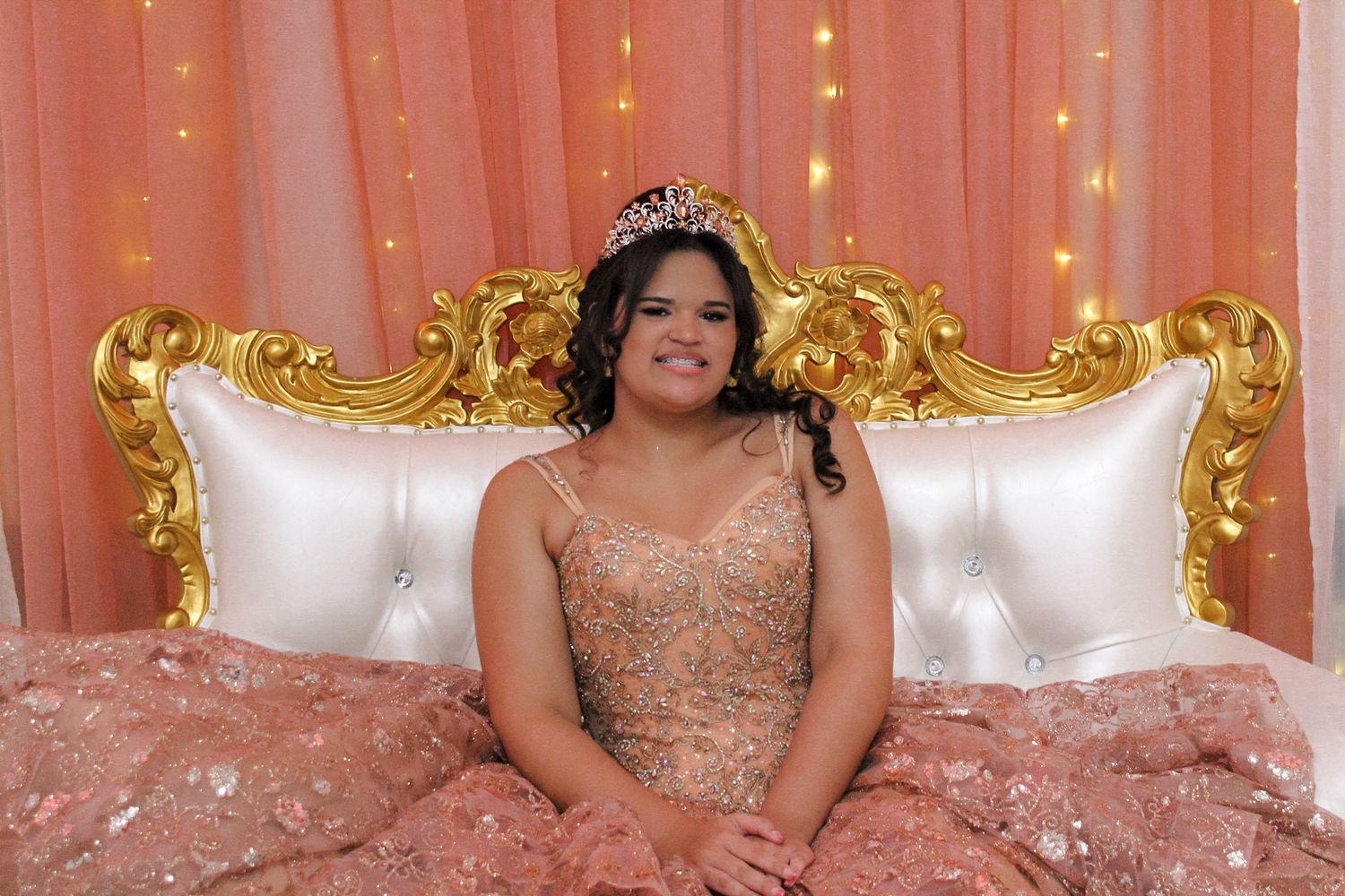 Quinceañera celebration portrait features guest of honor in rose gold gown on ornate white and gold throne.
