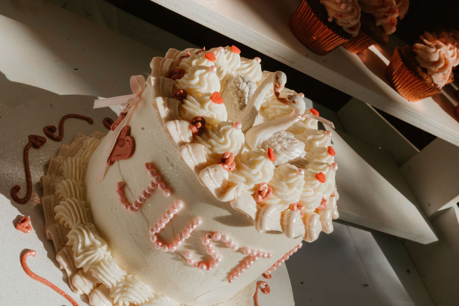Elegant white birthday cake with pink frosting decorations and festive cupcakes on a bright surface.