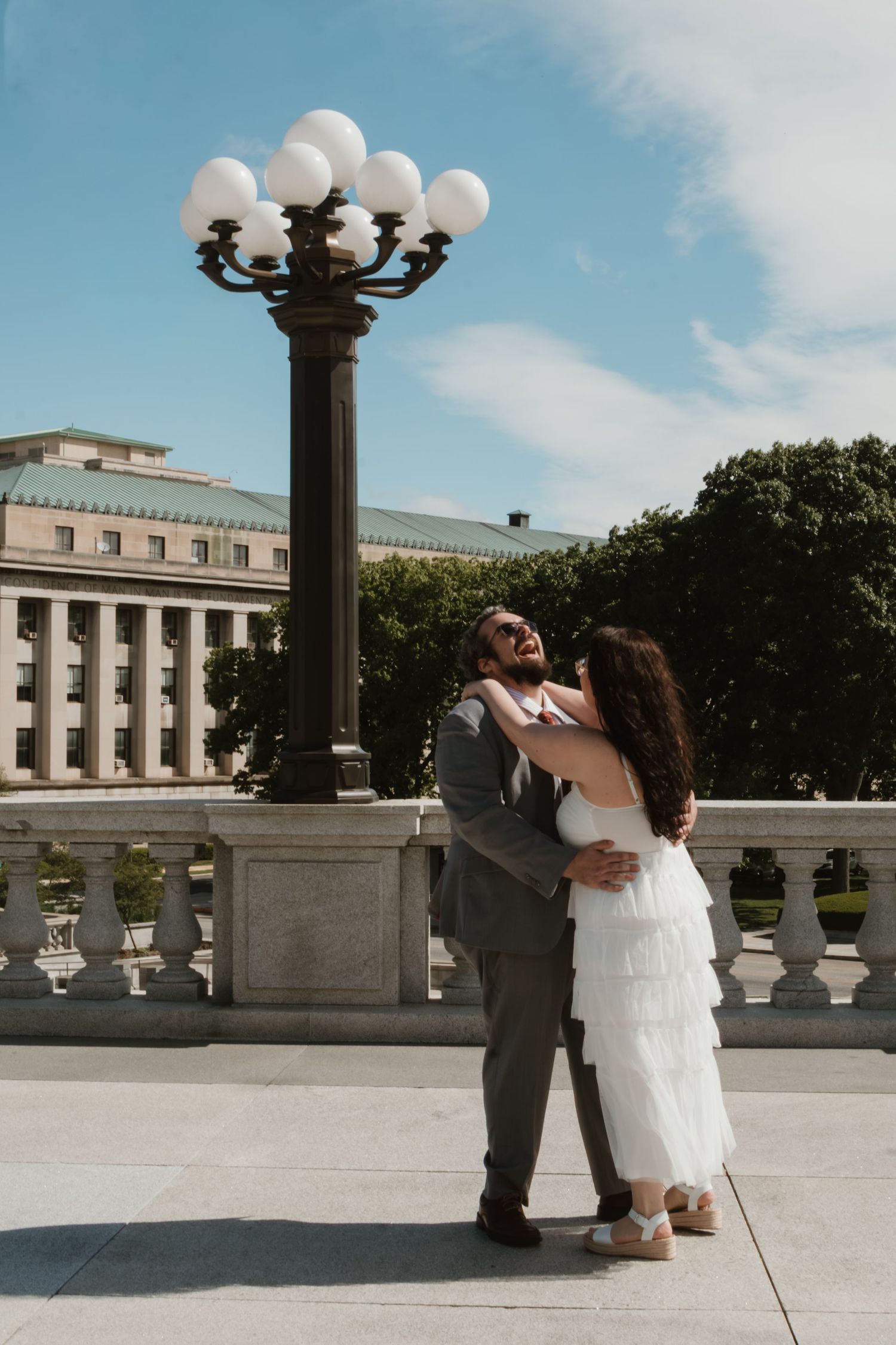 Couple embracing and laughing by ornate lamp post outside neoclassical building on sunny day.