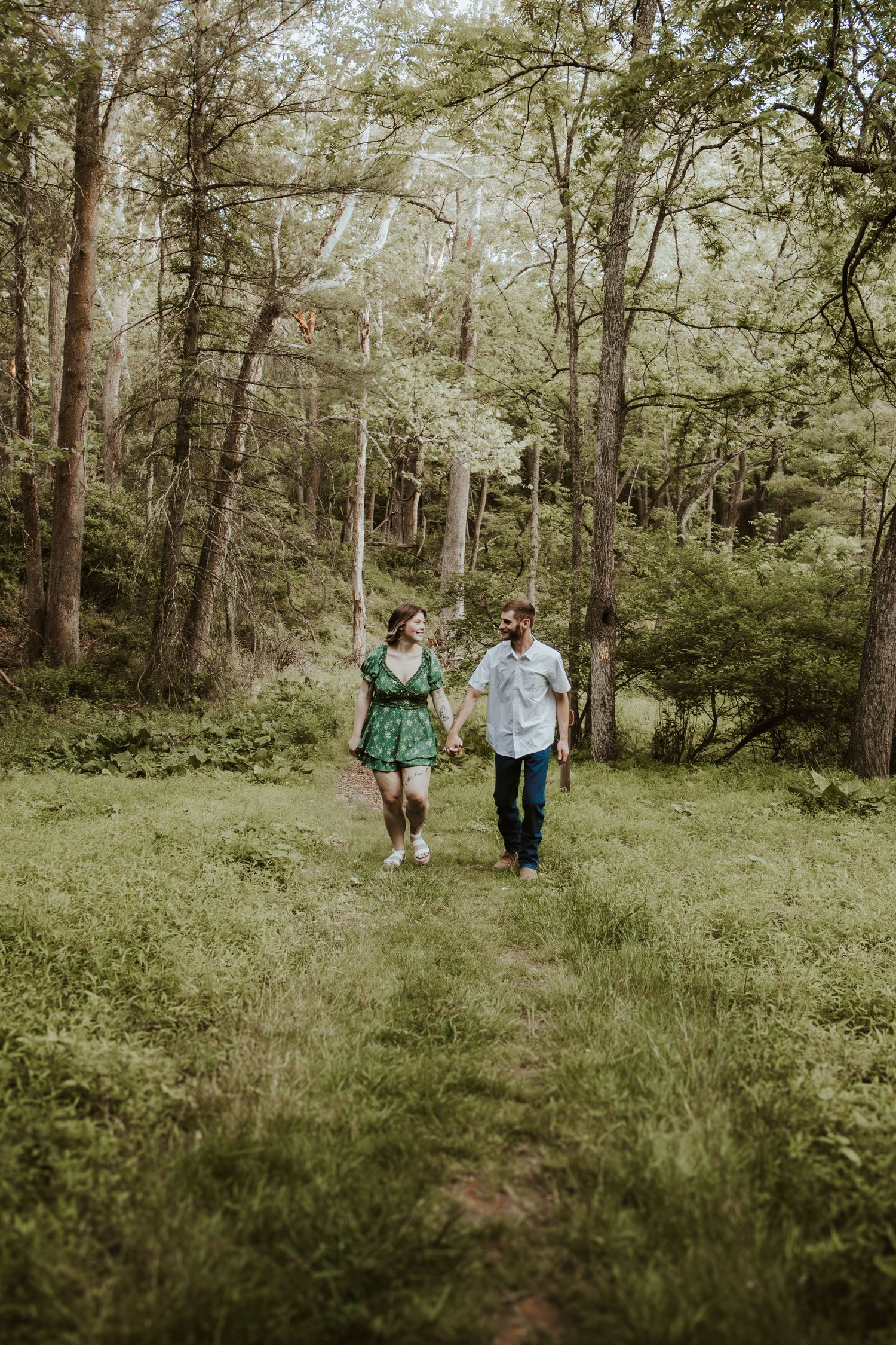 Couple holding hands walking on a forest path surrounded by lush green trees and grass.