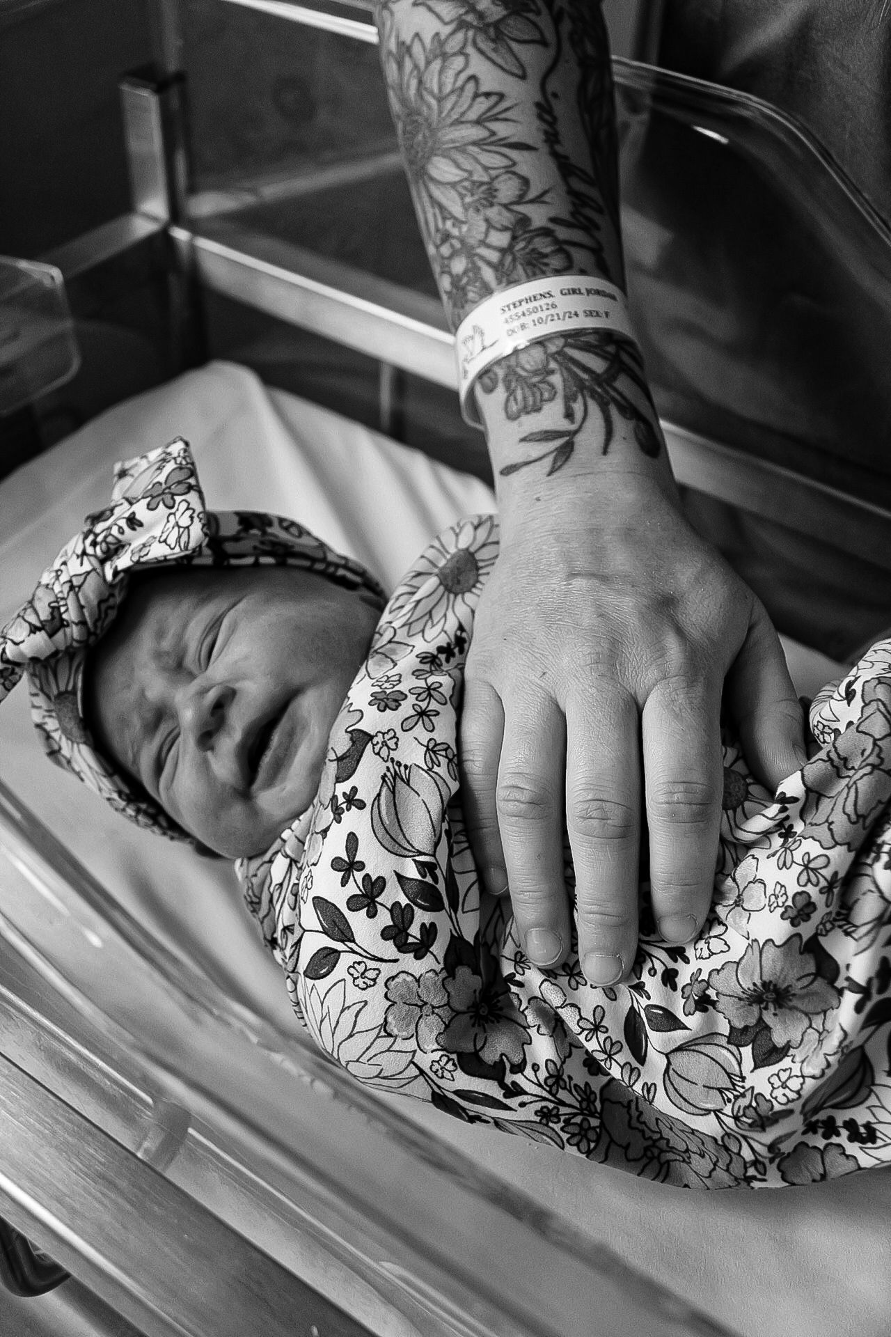 Black and white image of a tattooed hand resting on a swaddled newborn in a hospital bed.