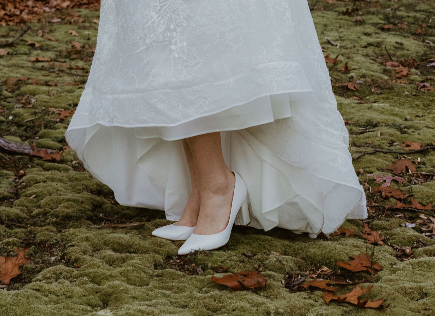 White wedding dress details showing the lace hem and bare feet on moss-covered stone with scattered fall leaves.