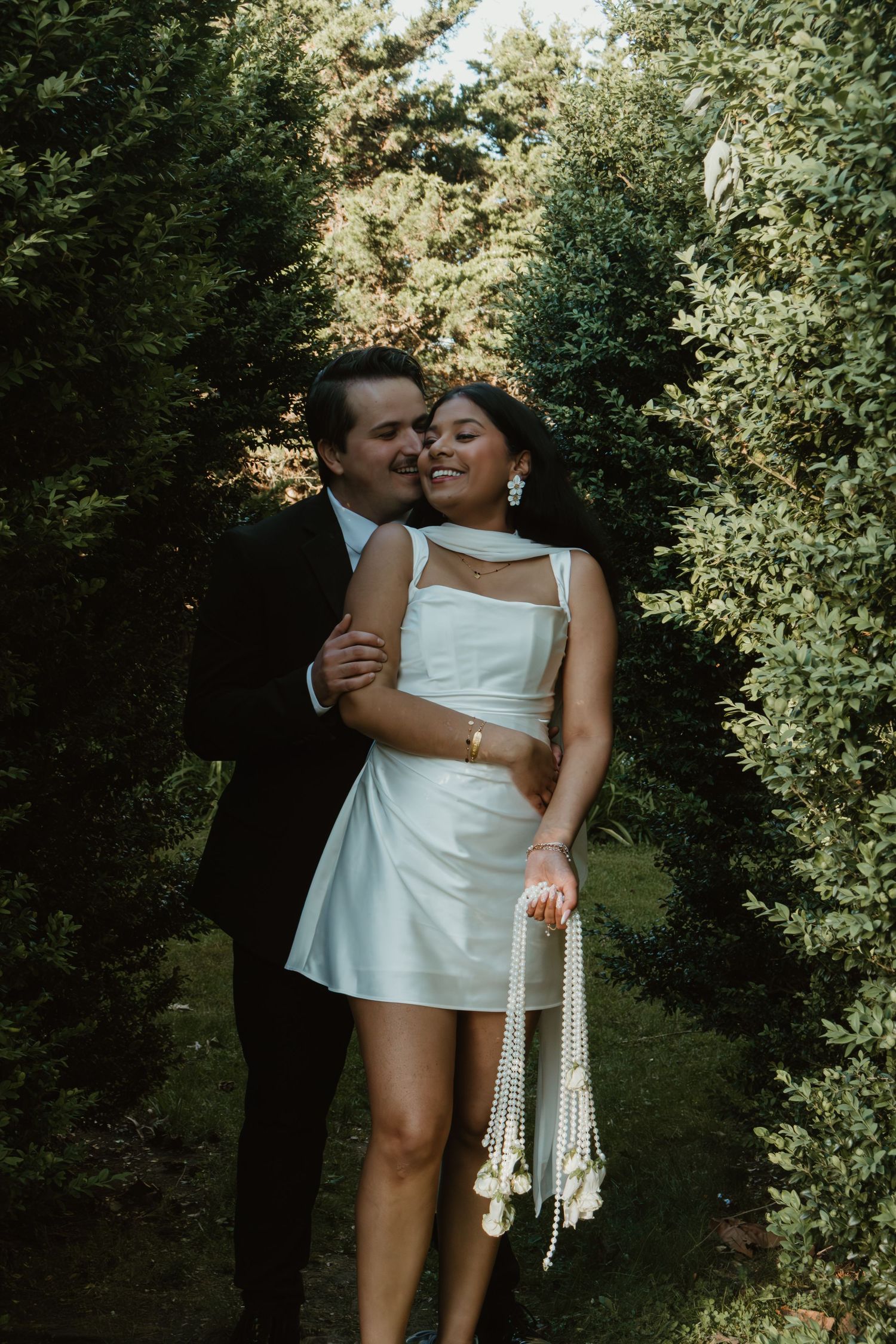 Couple embracing in garden, groom in black suit kissing bride wearing short white dress holding pearl accessories.