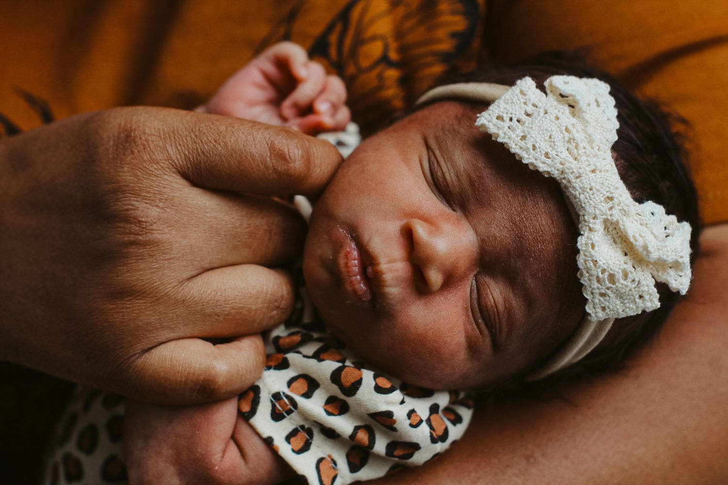 A sleeping infant wearing a lace headband and leopard onesie rests peacefully in mothers arms.