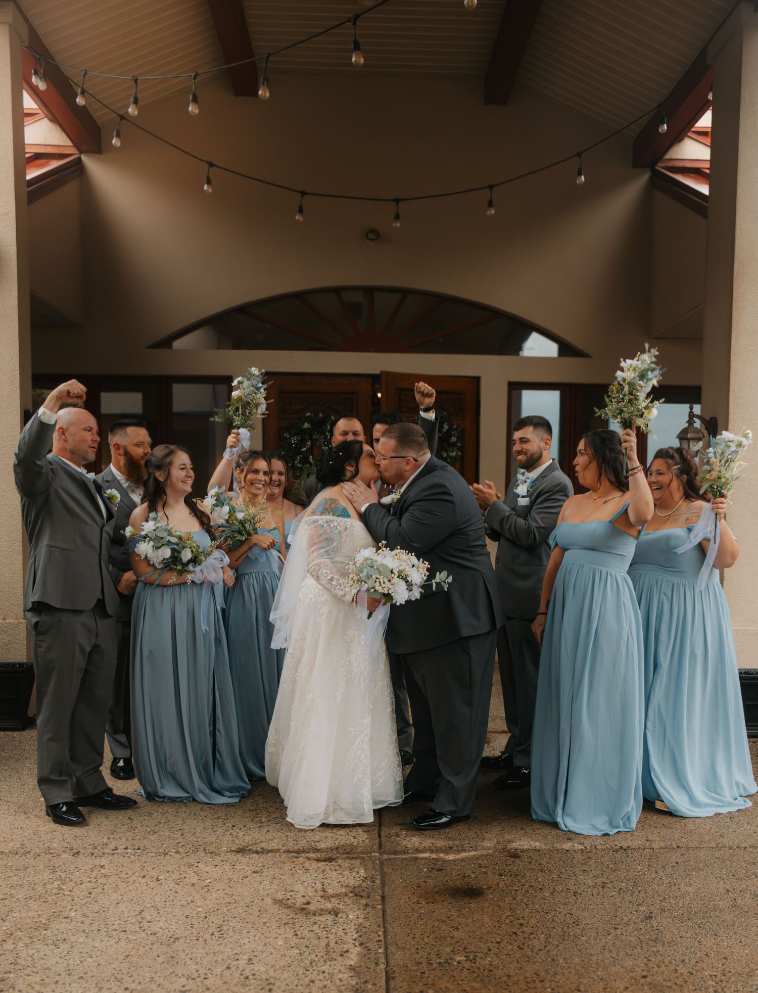Bride and groom share a kiss surrounded by wedding party in dusty blue gowns and gray suits outdoors.