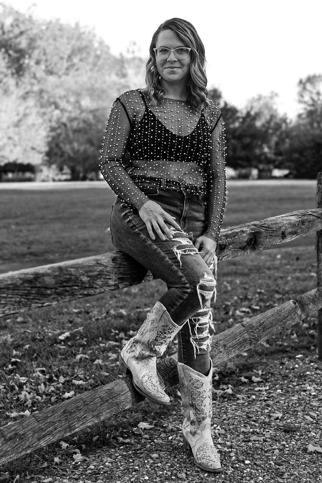 Black and white portrait featuring distressed jeans and sparkly top while sitting on rustic wooden fence.