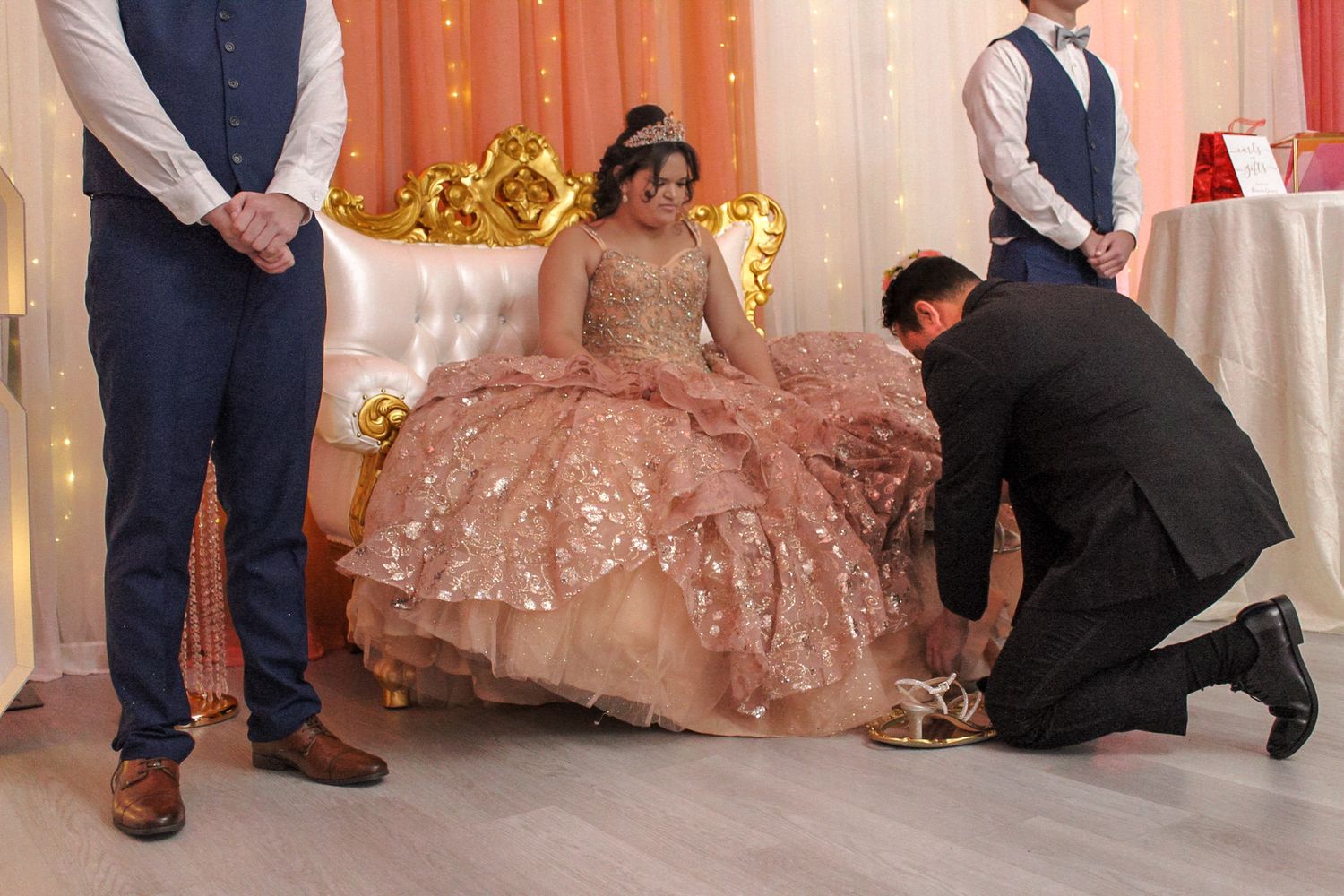 Person in a sparkly rose gold ballgown sits on ornate throne while attendants adjust the dress.