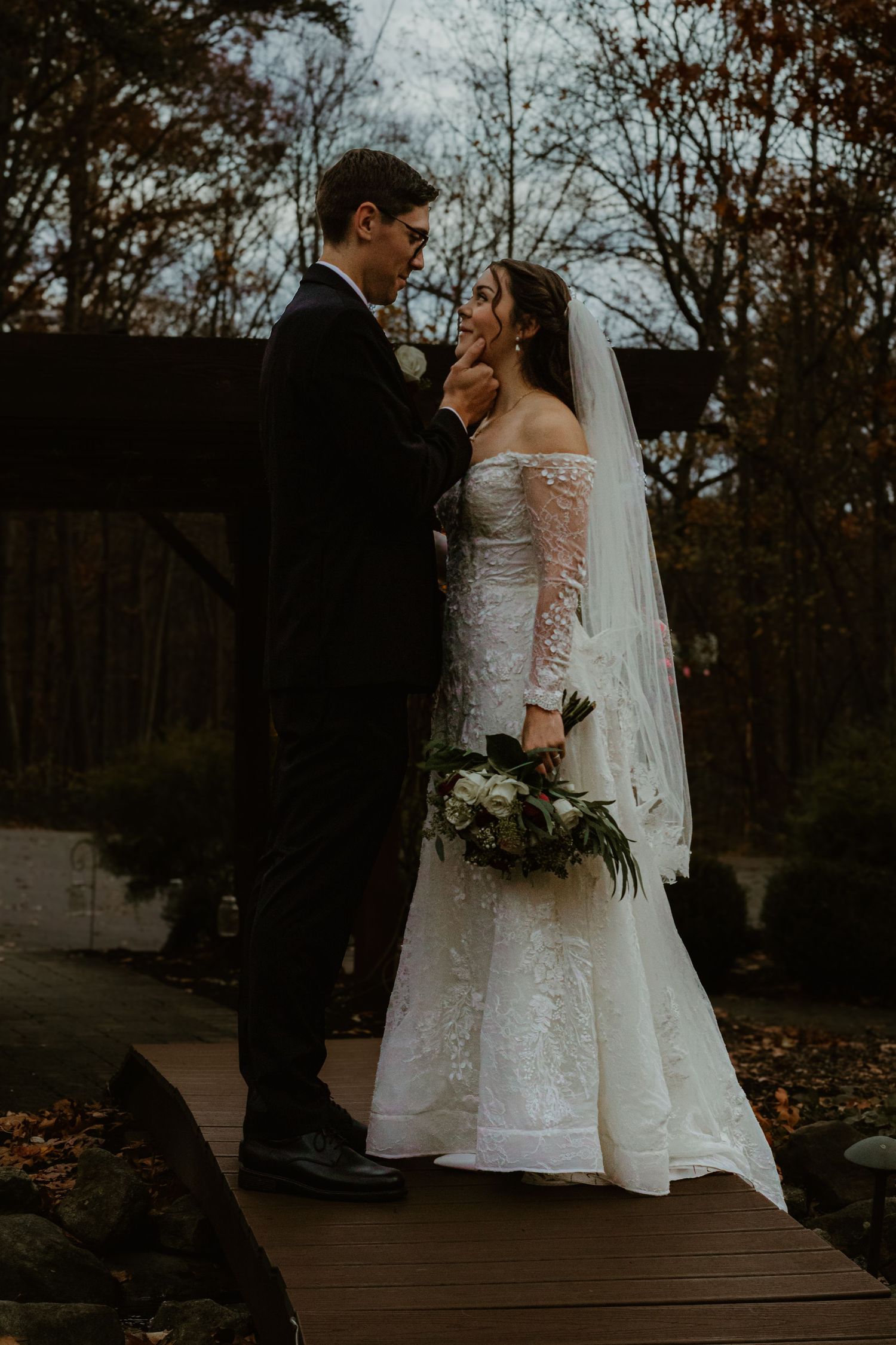 Wedding couple poses romantically on a wooden boardwalk surrounded by fall foliage at dusk.