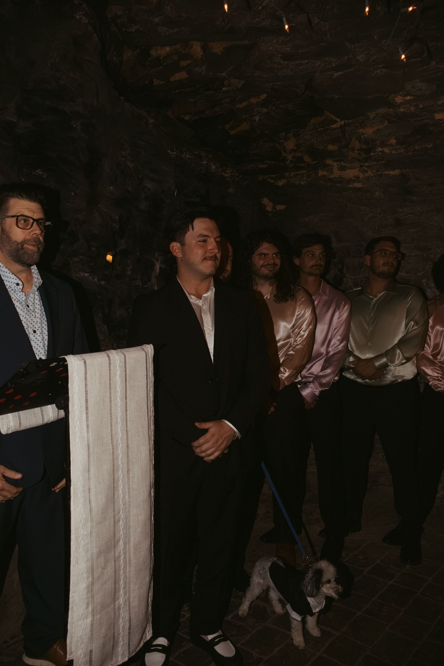 Group of men in formal attire standing in a dimly lit stone venue with a small dog on a leash.
