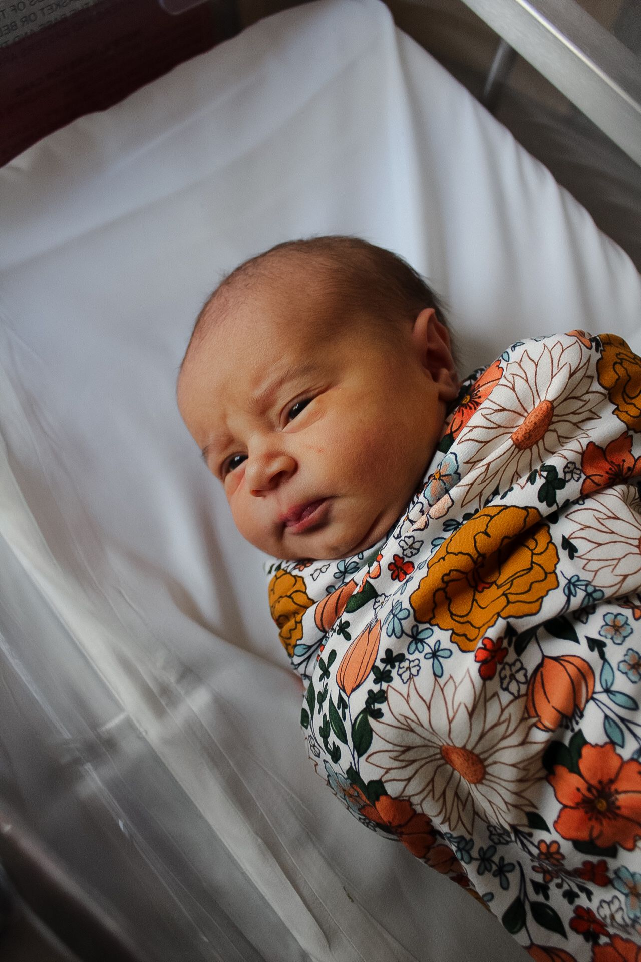Newborn baby wearing a floral printed swaddle sleeps peacefully in a hospital bassinet.