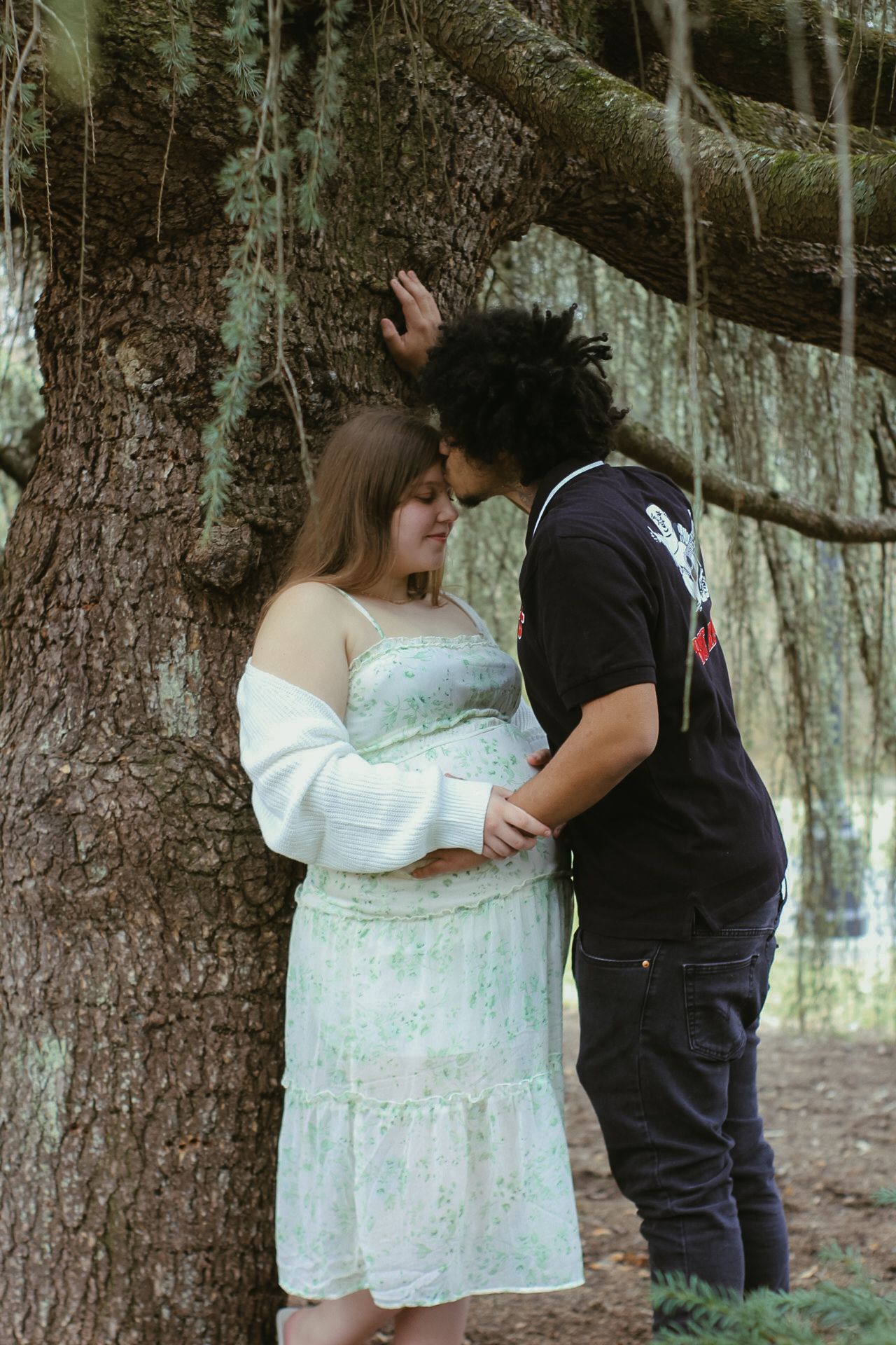 A couple shares an intimate moment in the woods against a large tree trunk.