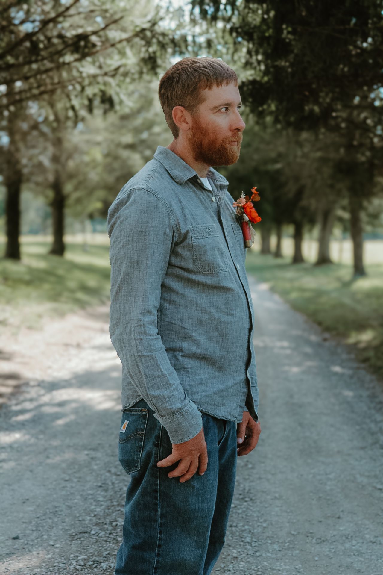 A groom in a grey suit jacket and jeans stands casually on a gravel path surrounded by evergreen trees.