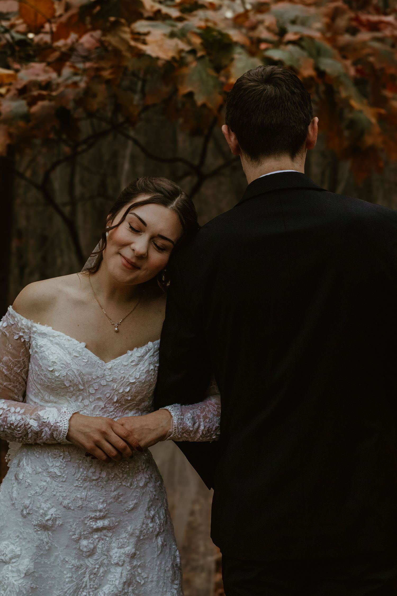 Bride in delicate lace wedding dress poses gracefully against autumn foliage in a romantic woodland setting.