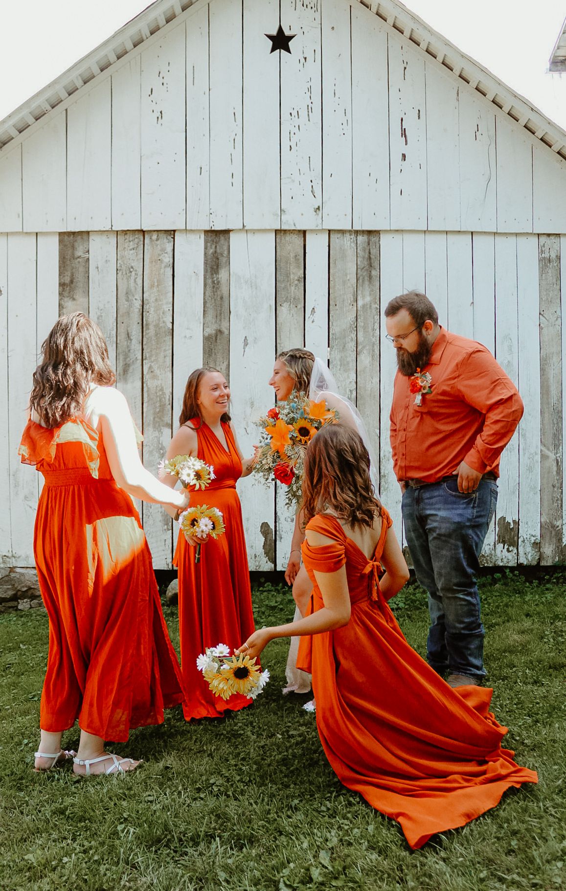 Group in vibrant orange dresses gather for photos in front of a rustic white barn on a grassy lawn.