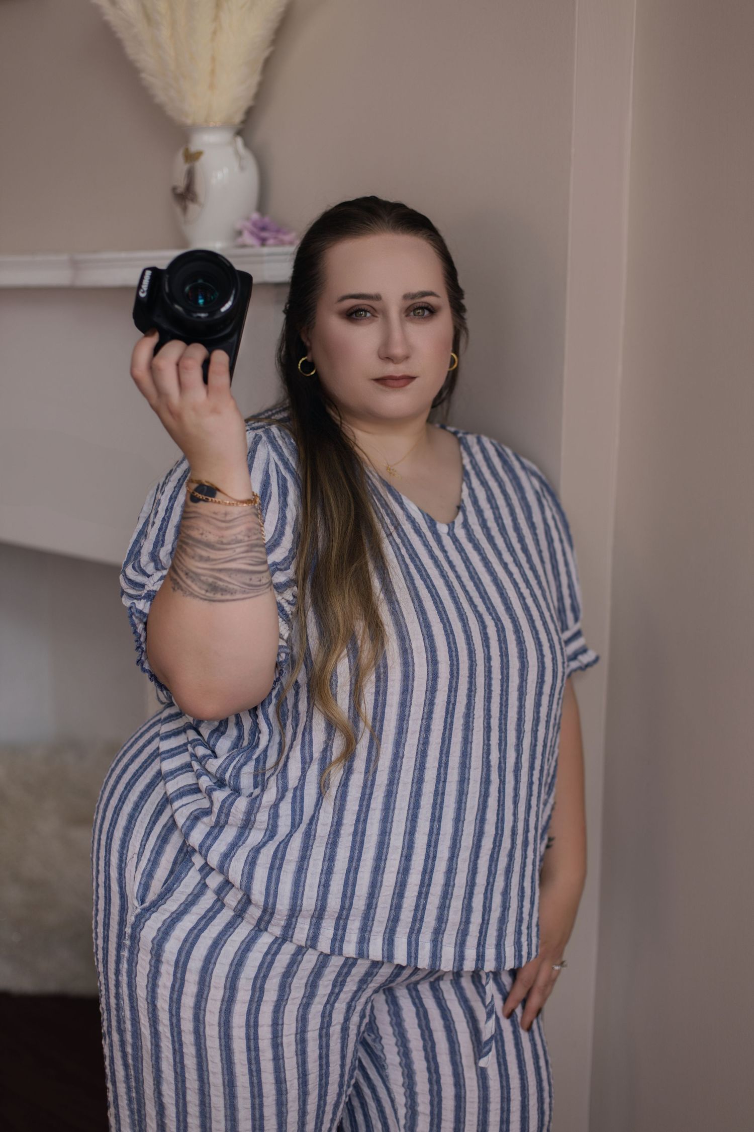Portrait of photographer in striped outfit holding camera against neutral wall with decorative shelf.
