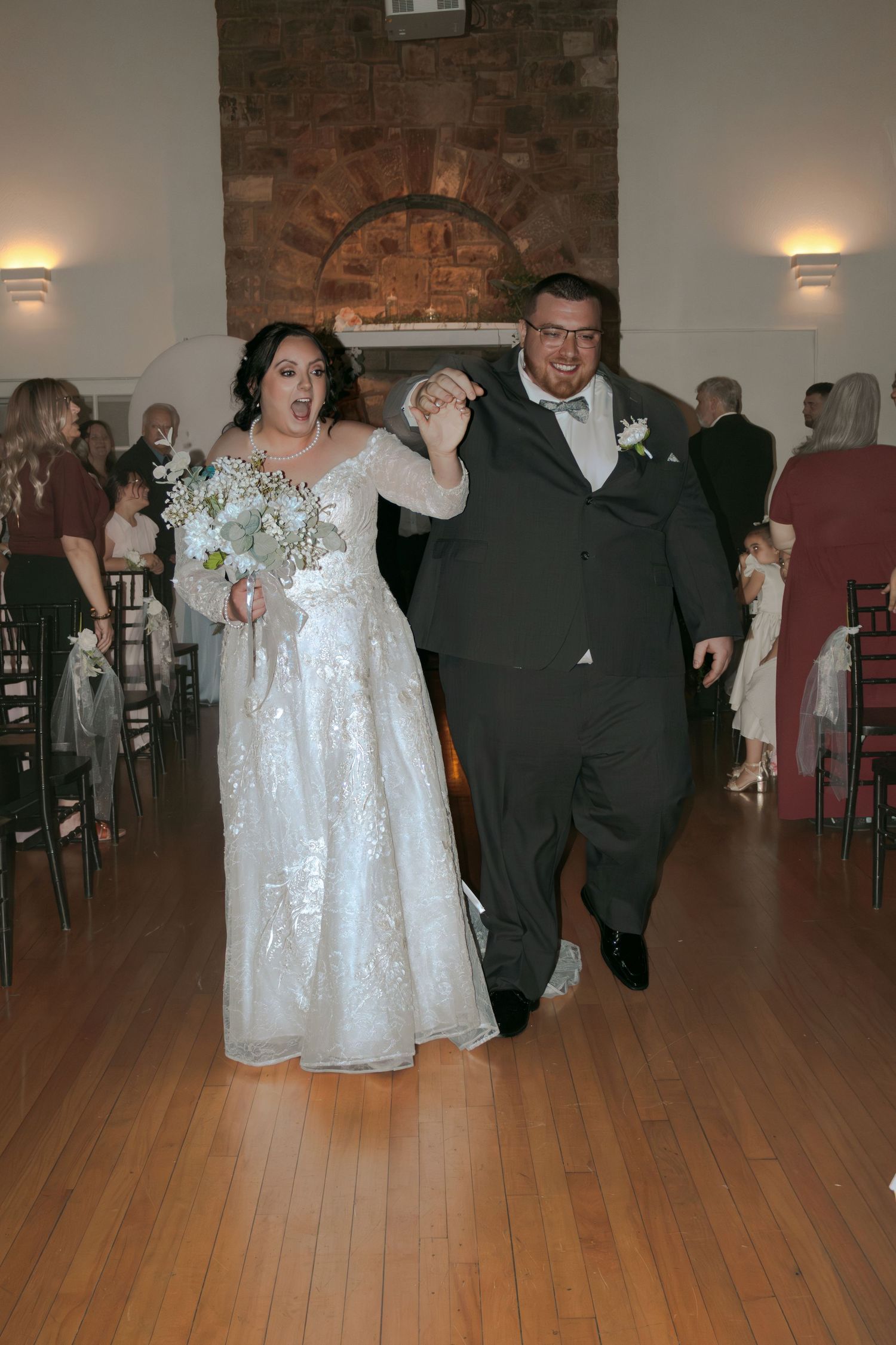 Bride in white lace gown and groom in gray suit walk down aisle holding hands at elegant indoor wedding venue.