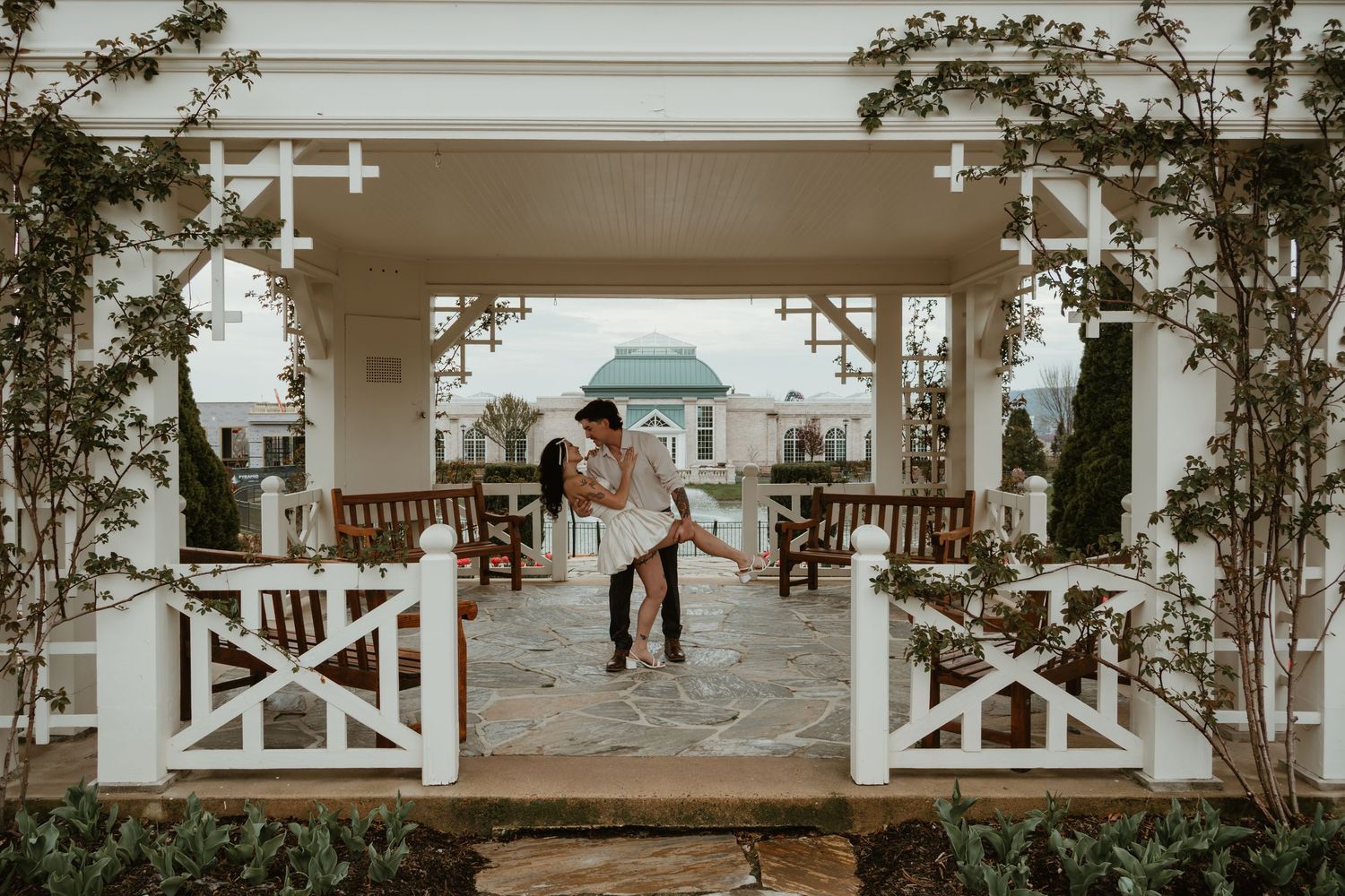 Couple dancing romantically under a white pergola surrounded by lush greenery at an elegant outdoor venue.
