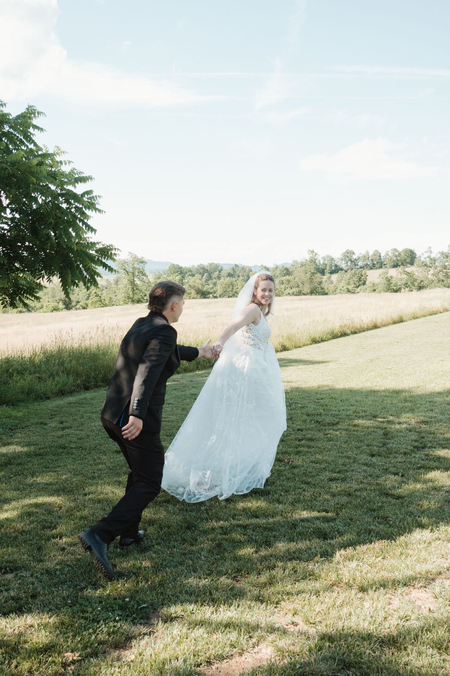 Bride and groom holding hands running through green meadow on sunny wedding day.