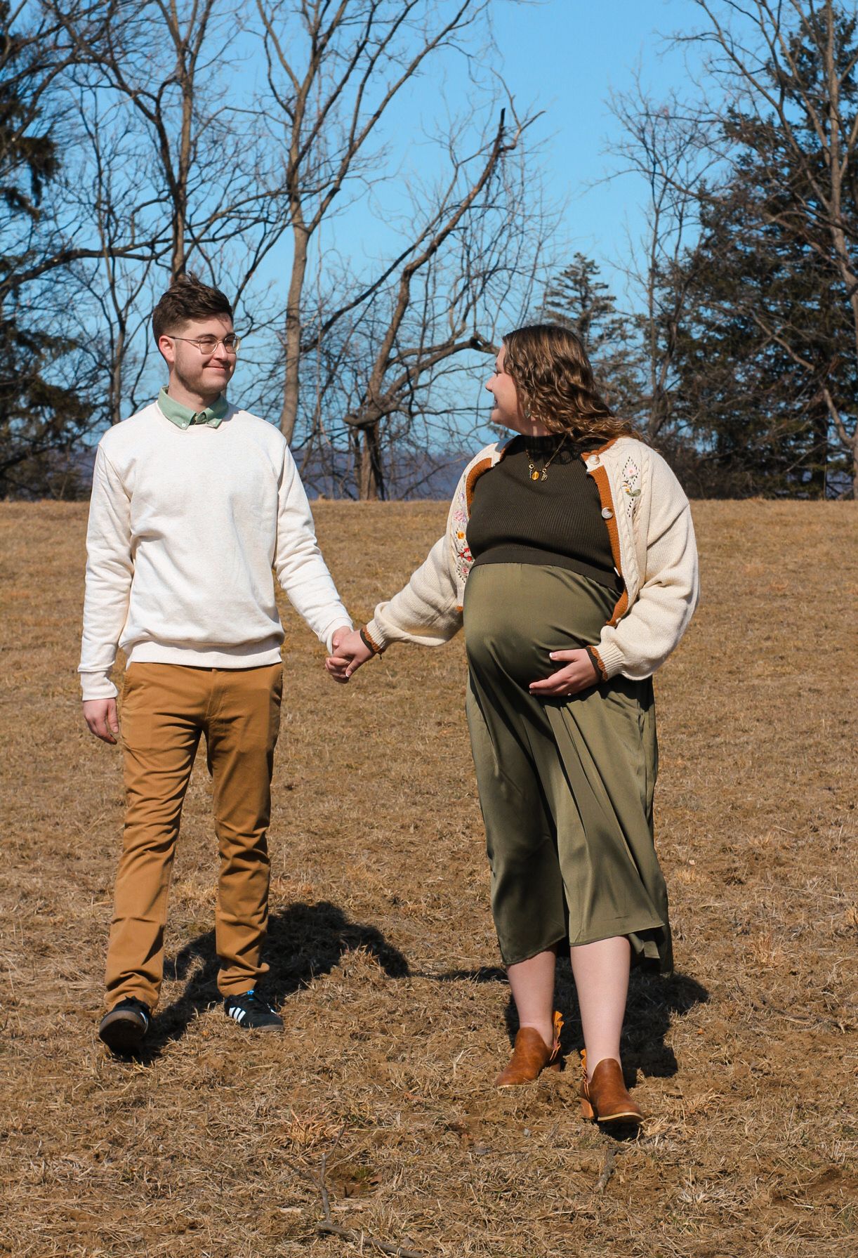 Couple holding hands in a sunny field with bare trees in background.