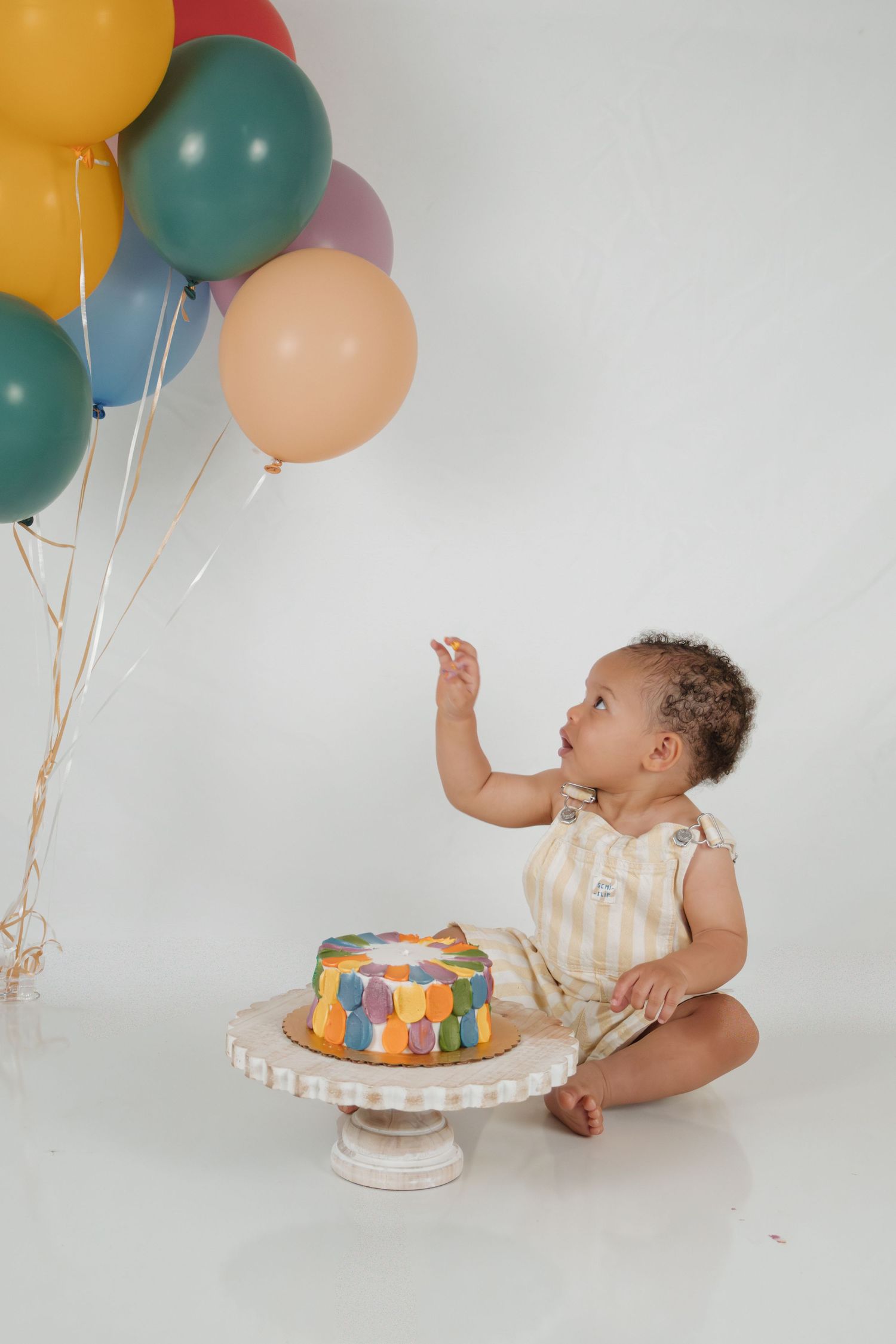 Baby's first birthday cake smash with colorful balloons and a decorated cake on a white cake stand.