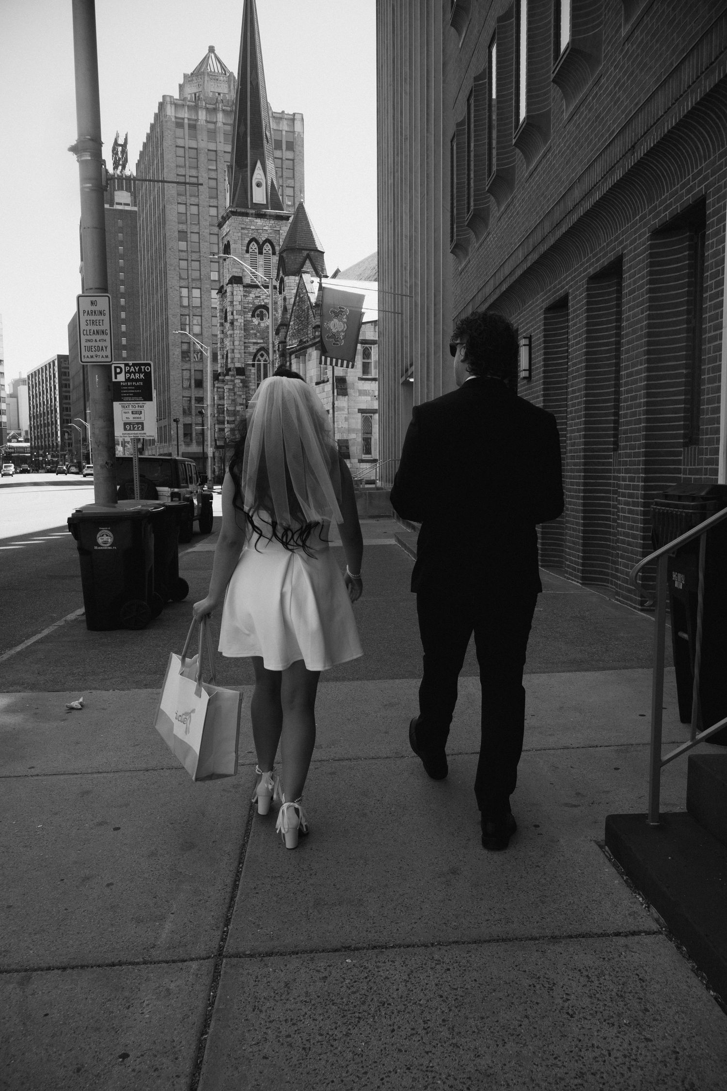 Black and white photo of a bride and groom walking down a city sidewalk past tall buildings.