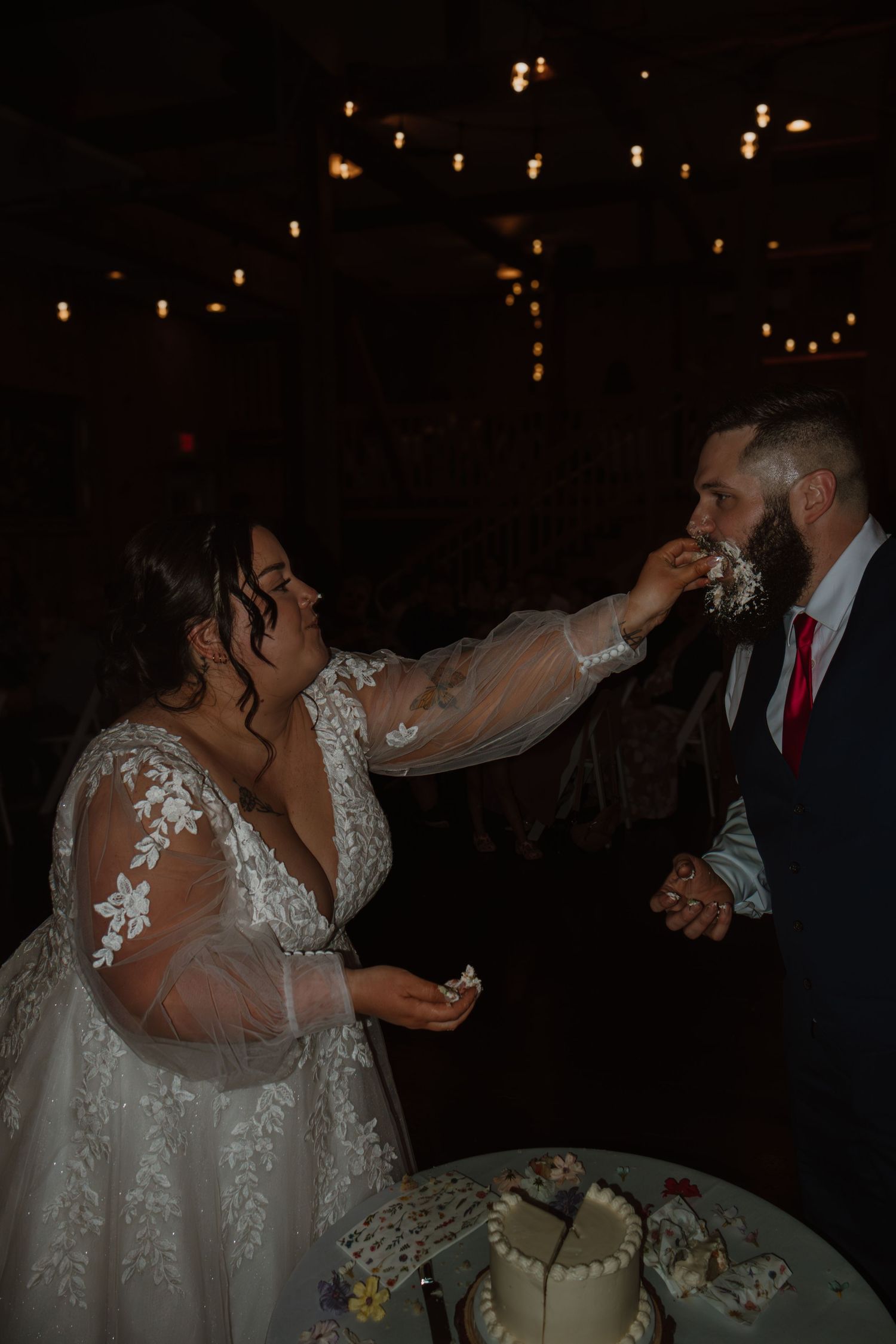 Bride smashing wedding cake into groom's face during reception in dimly lit venue with string lights.