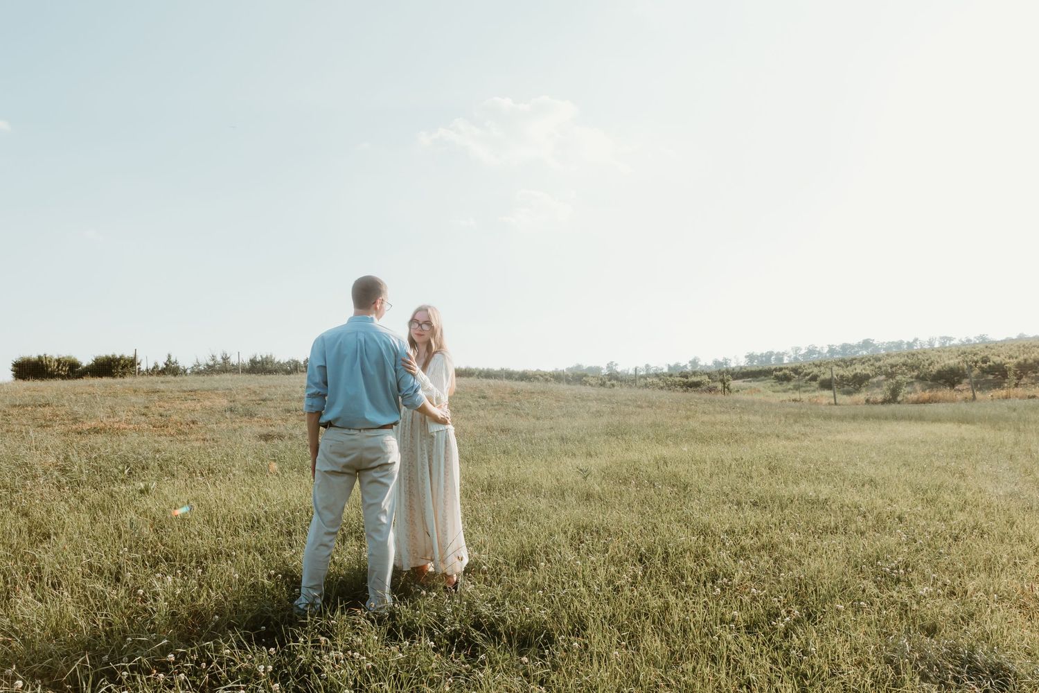Couple holding hands in a lush green meadow on a sunny day, woman in white dress, man in blue shirt.