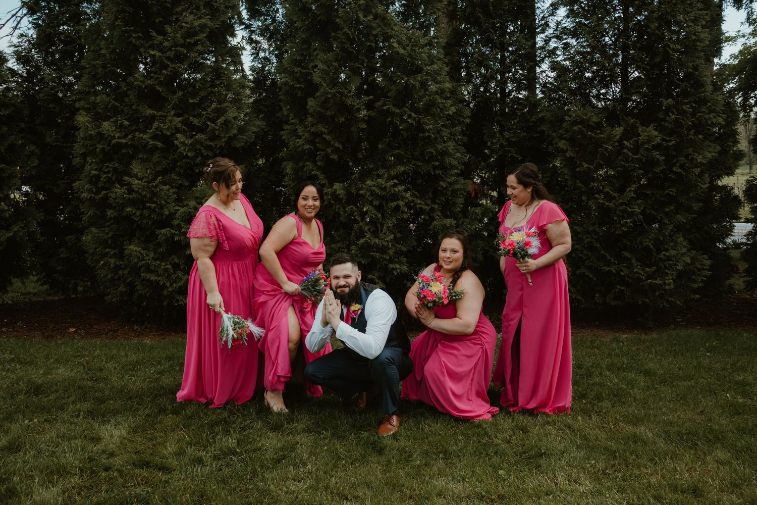 Groom crouching with four bridesmaids in pink dresses posing outdoors on green grass with evergreen trees.