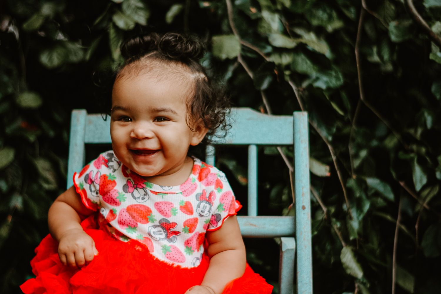 Infant wearing red outfit smiling brightly while seated outdoors.