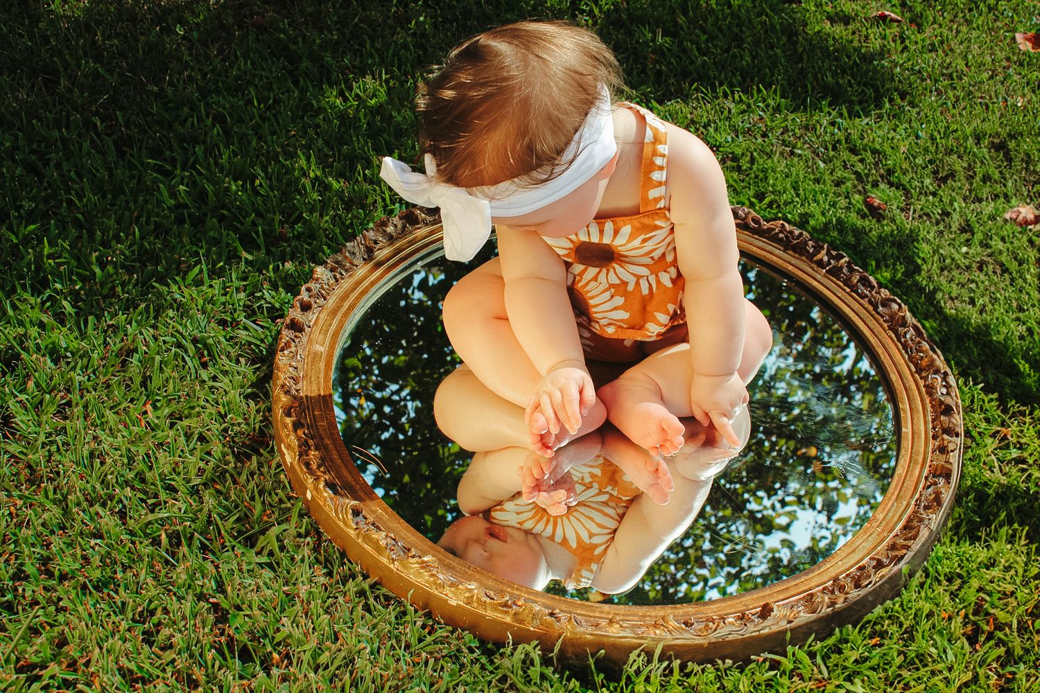 A child sits on a decorative mirror placed on grass, creating a whimsical reflection.