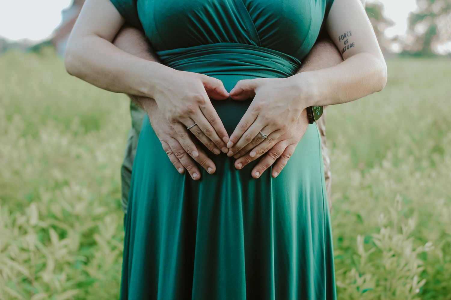 Hands forming a heart shape on a pregnant belly in a green dress against a grassy background.