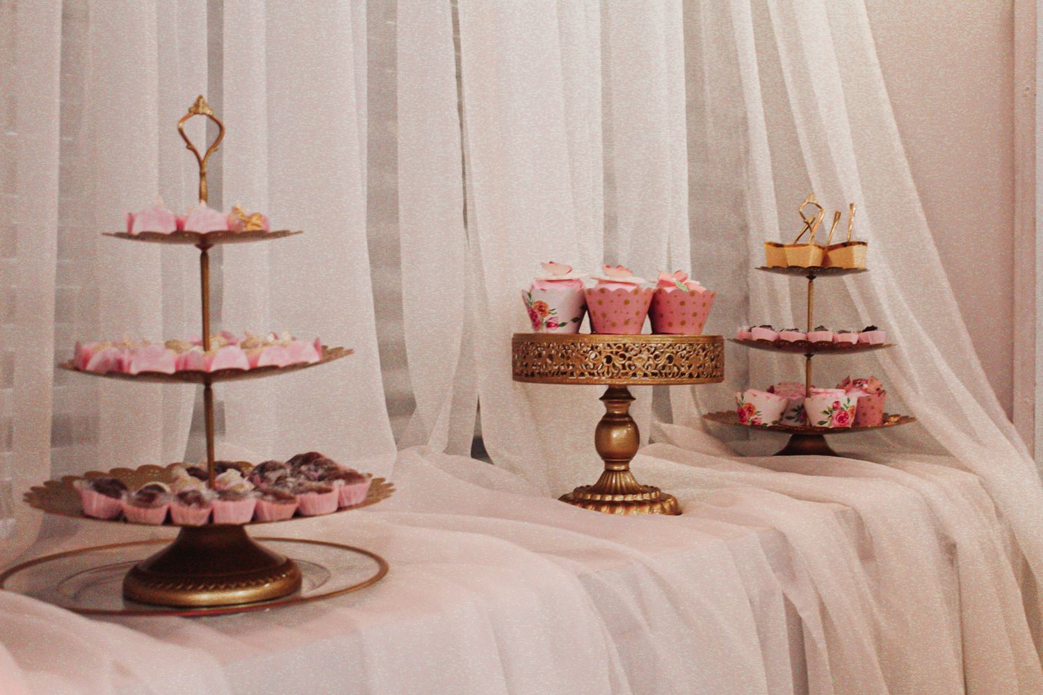Vintage gold tiered stands displaying pink cupcakes and desserts on white tablecloth.