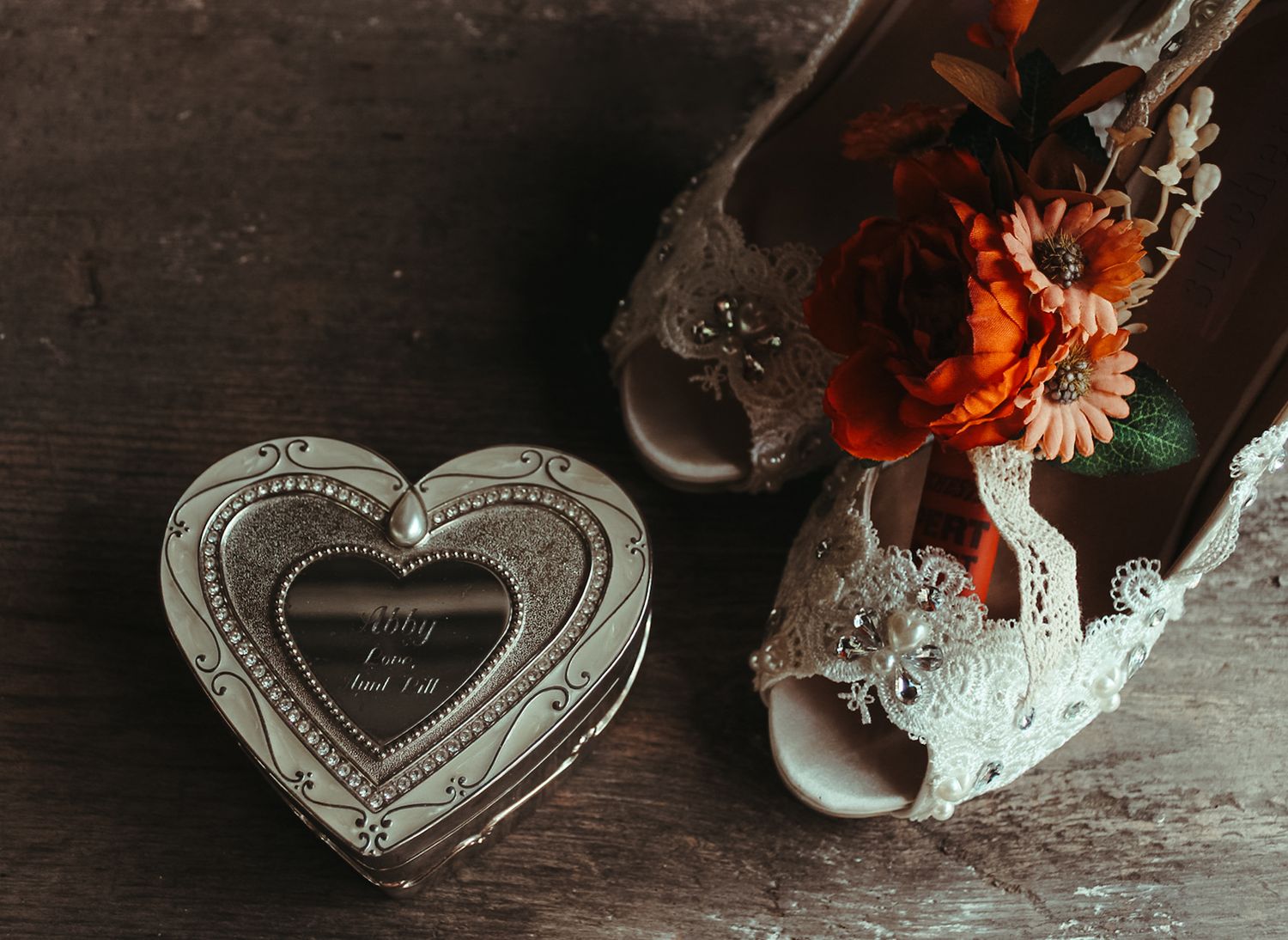 Vintage silver heart-shaped jewelry box beside white ceramic vase with orange flowers on dark wooden surface.