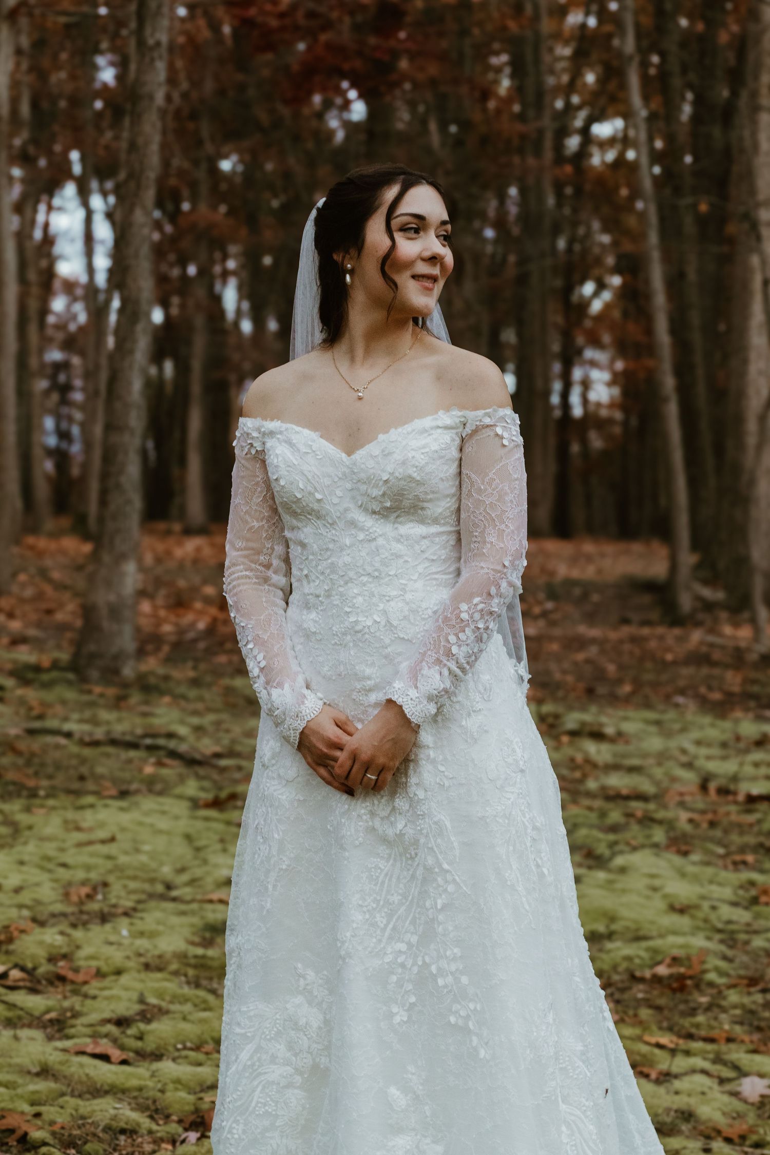 Full-length view of an elegant white wedding gown with long lace sleeves in an autumn forest setting.