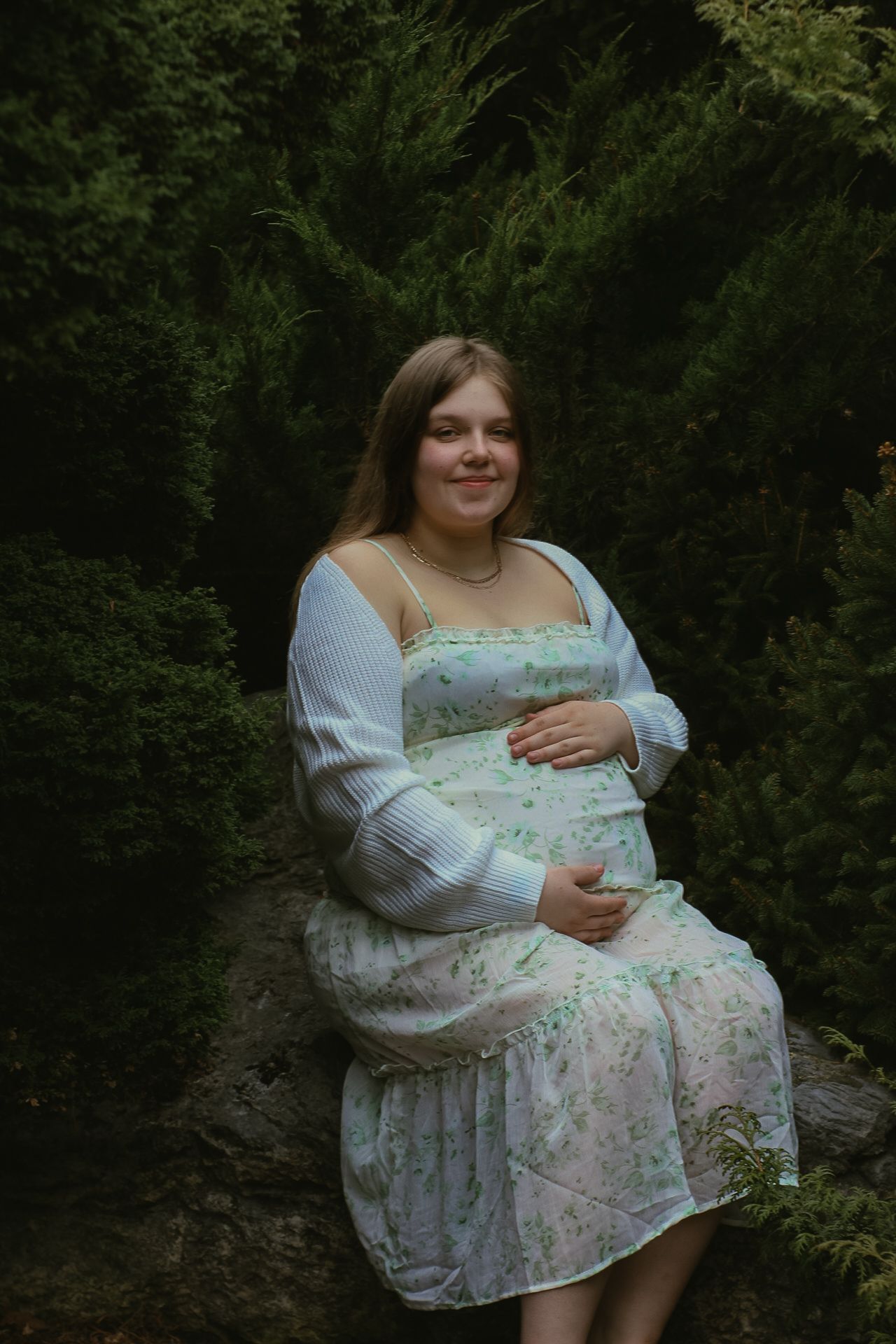 An expecting mother in a floral dress sits on a rock surrounded by lush greenery.