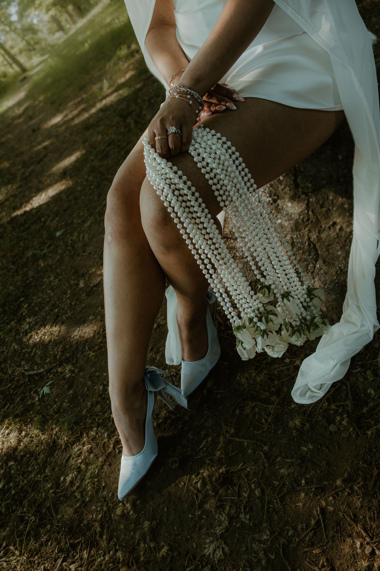Bride in white dress holding pearl bouquet with blue heels sitting outdoors in dappled sunlight.