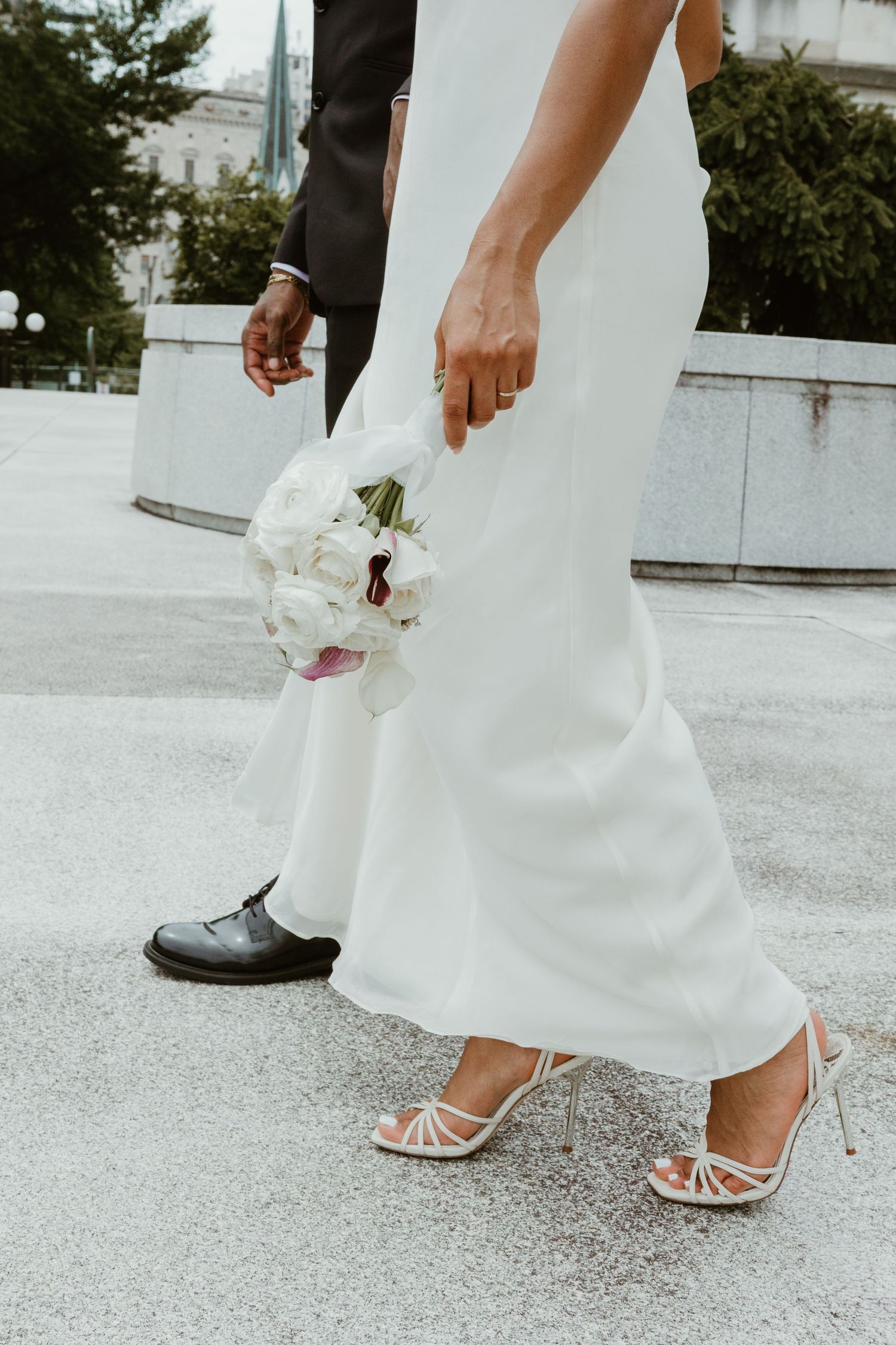 Bride in white gown and strappy heels holding white bouquet beside groom in black suit on wedding day.