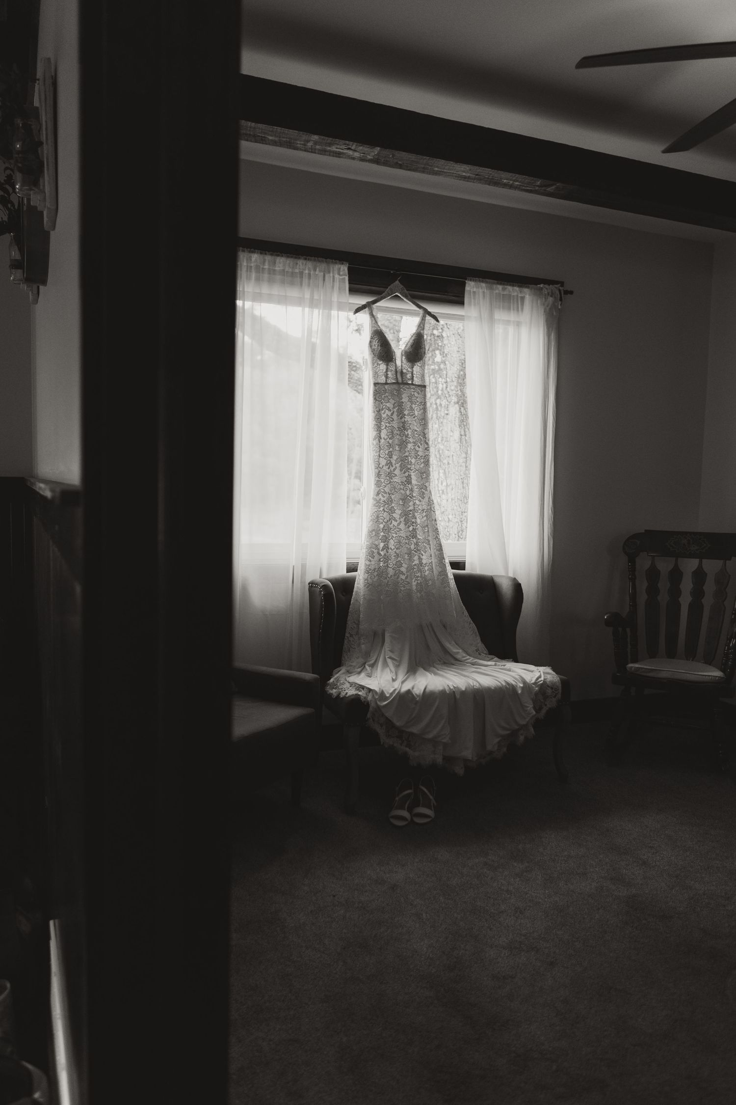 Black and white photo of an elegant wedding dress hanging by a window, with a long lace train draped over a chair.