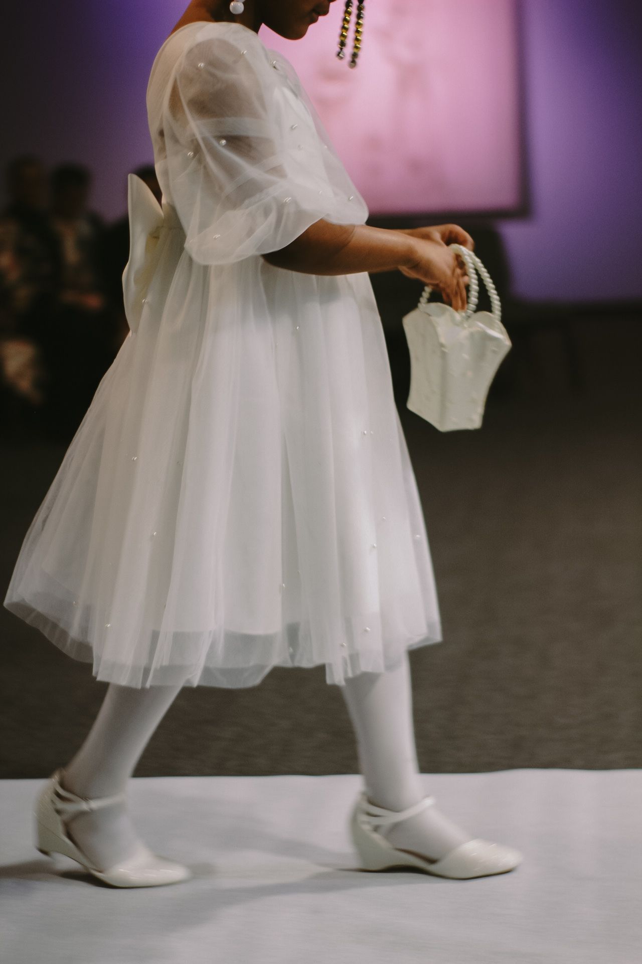 Child in white tulle dress and matching shoes walking across stage during formal event.