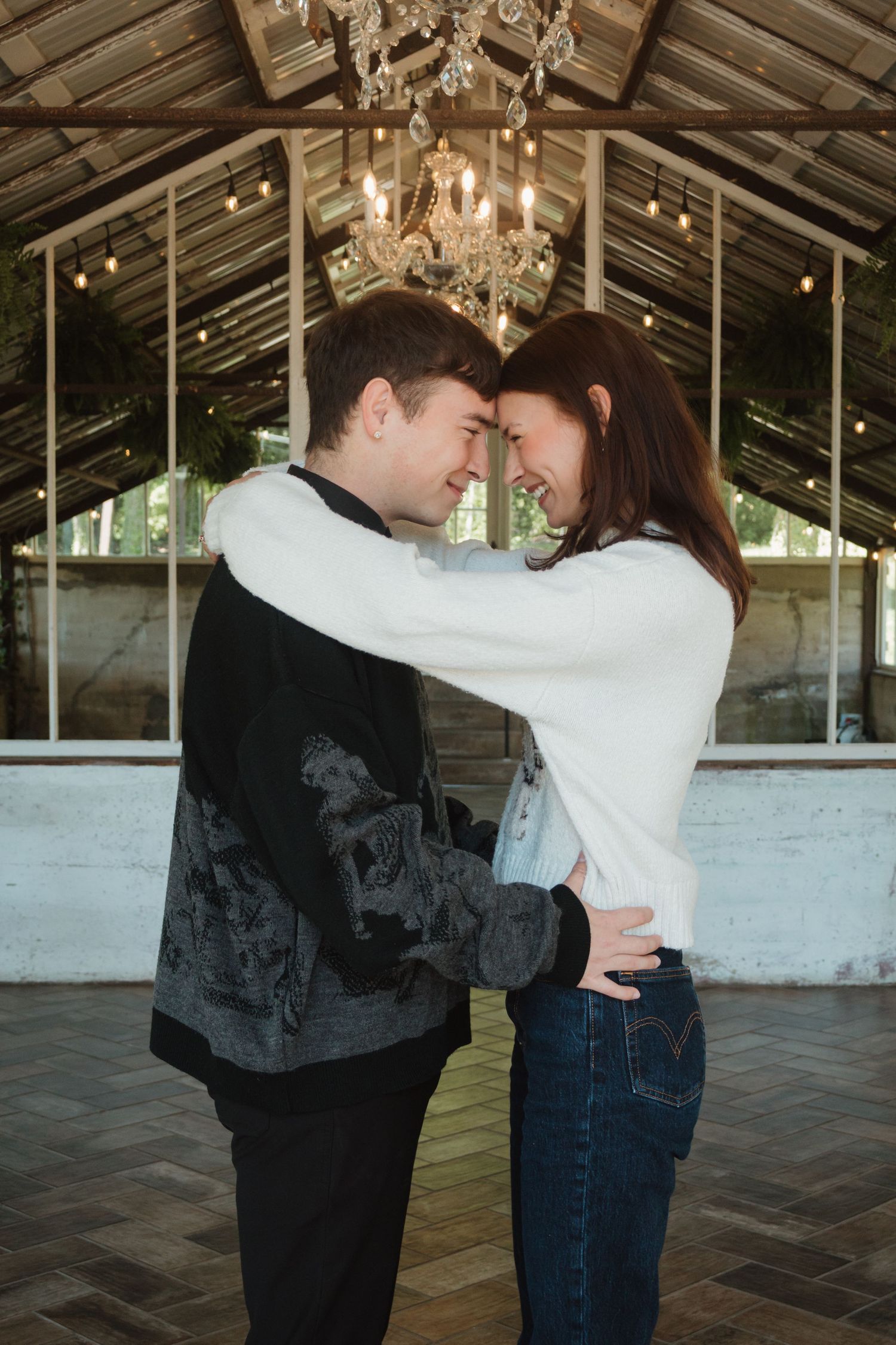 A young couple embraces and smiles at each other inside a rustic greenhouse venue with a chandelier overhead.