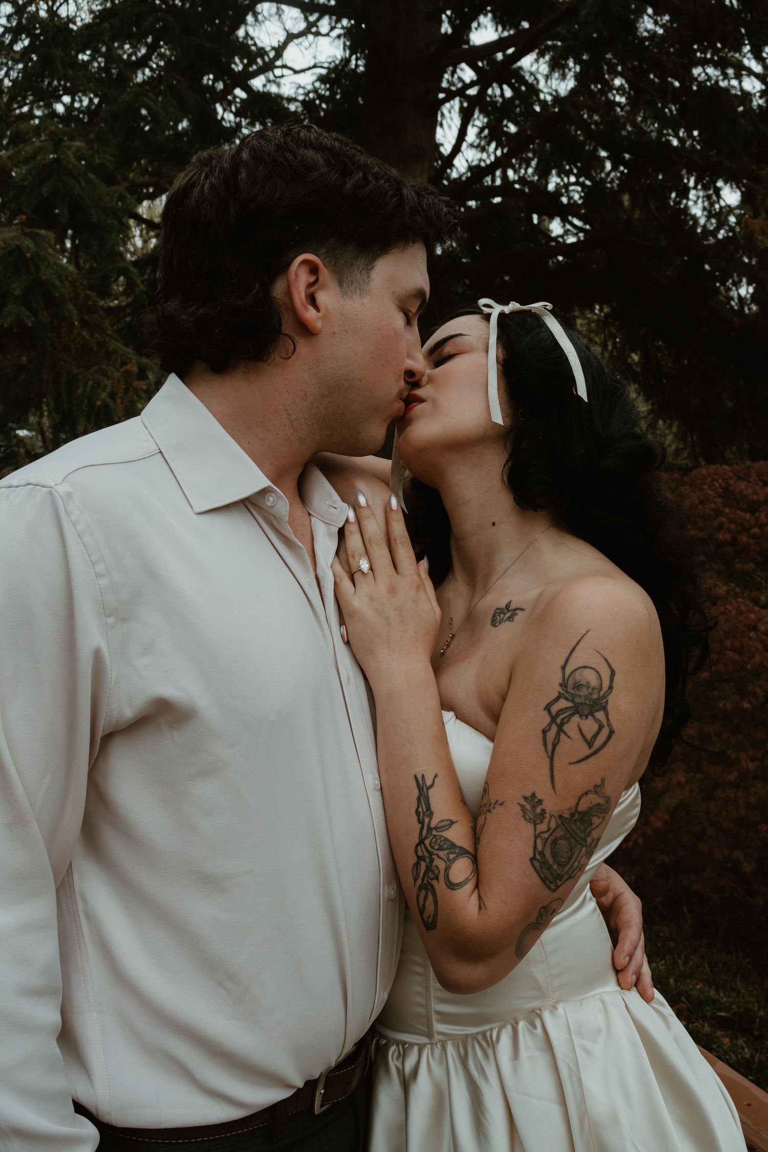 A tattooed bride in a white strapless gown kisses her groom in a white shirt outdoors among lush green trees.