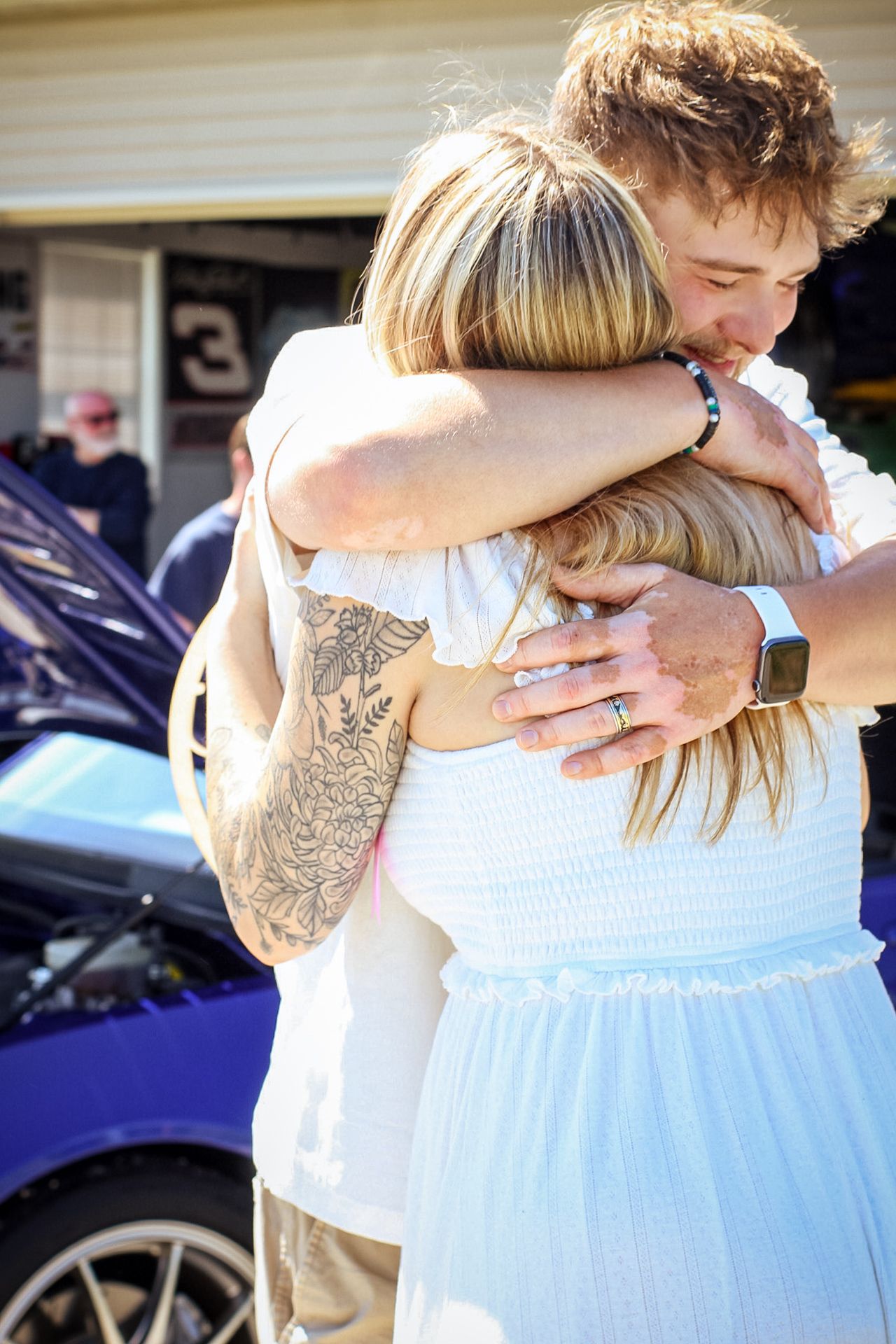 A tender embrace between two people, one with visible arm tattoos wearing a white shirt.