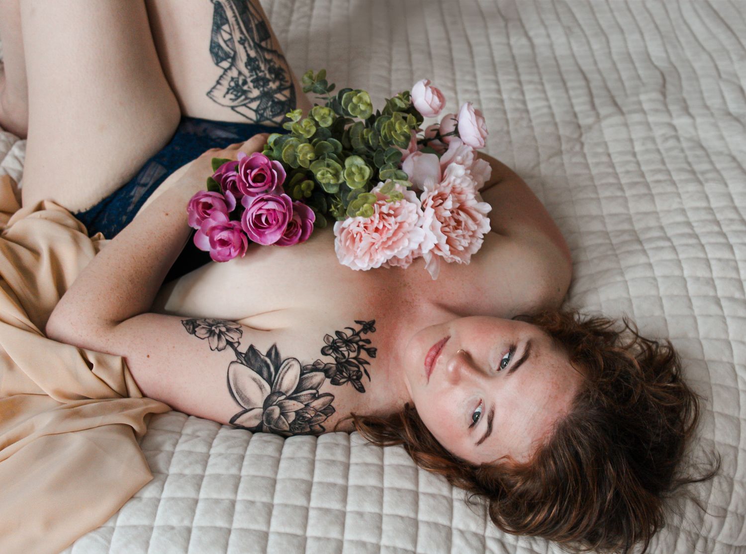 Woman lying on white bedding with scattered pink roses and greenery in romantic boudoir photography series.