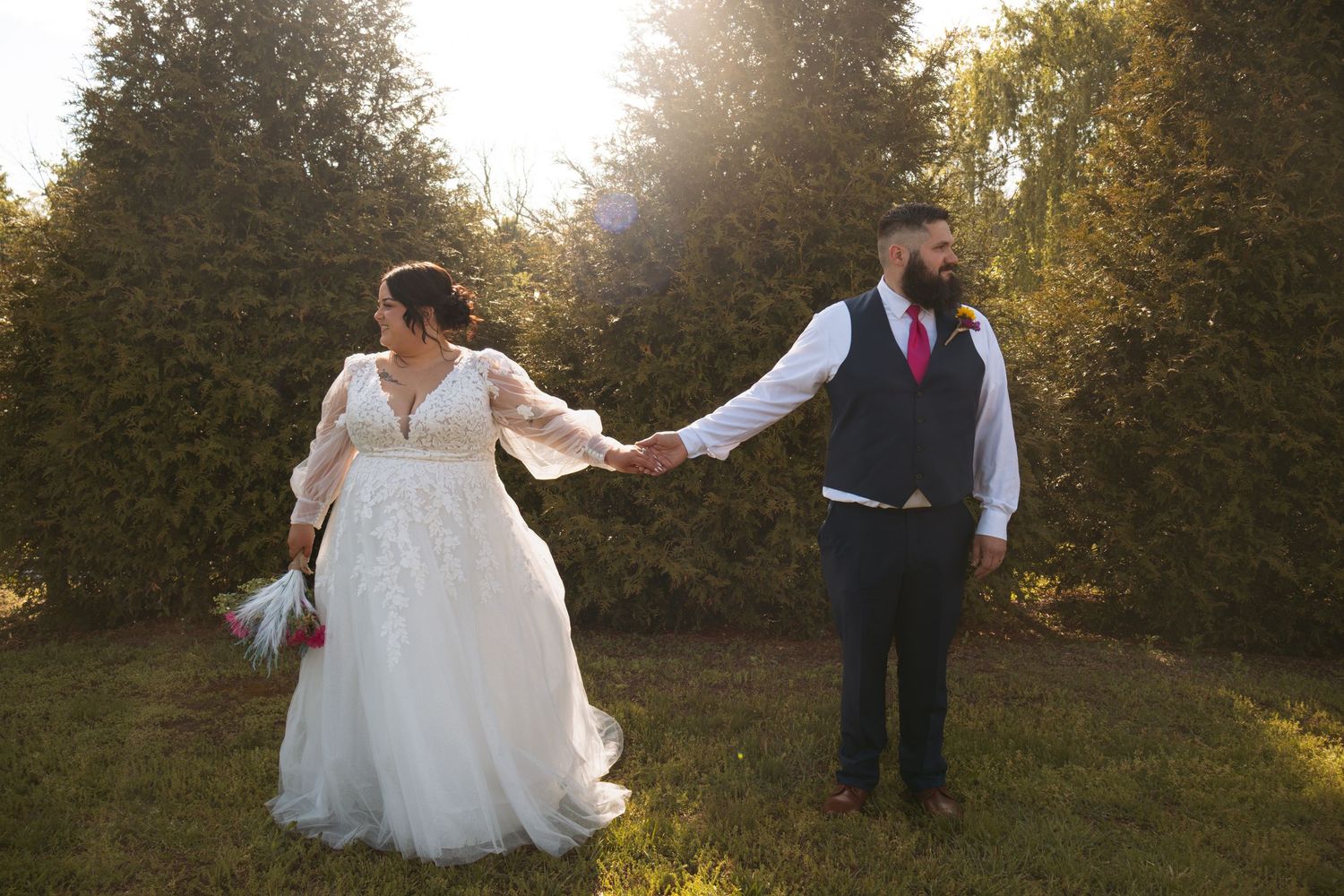 Bride in white lace gown holds groom's hand in navy suit with pink tie, posing in sunlit garden.