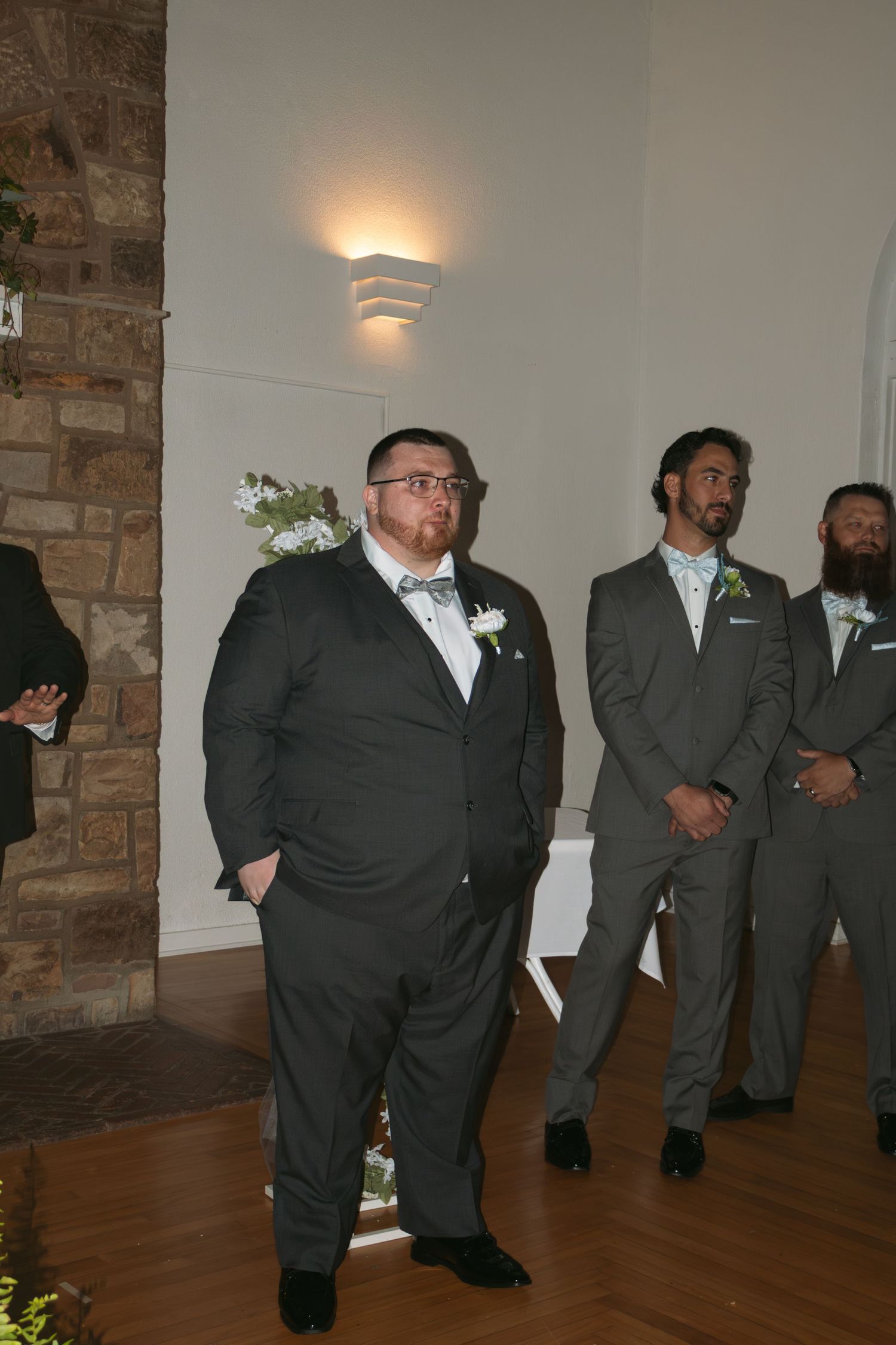 Groomsmen in gray suits with boutonnieres stand attentively during a wedding ceremony in an elegant indoor venue.