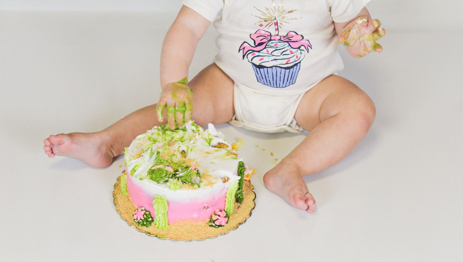 A cake smash photo featuring green frosting and tiny feet beside a decorated pink cake.