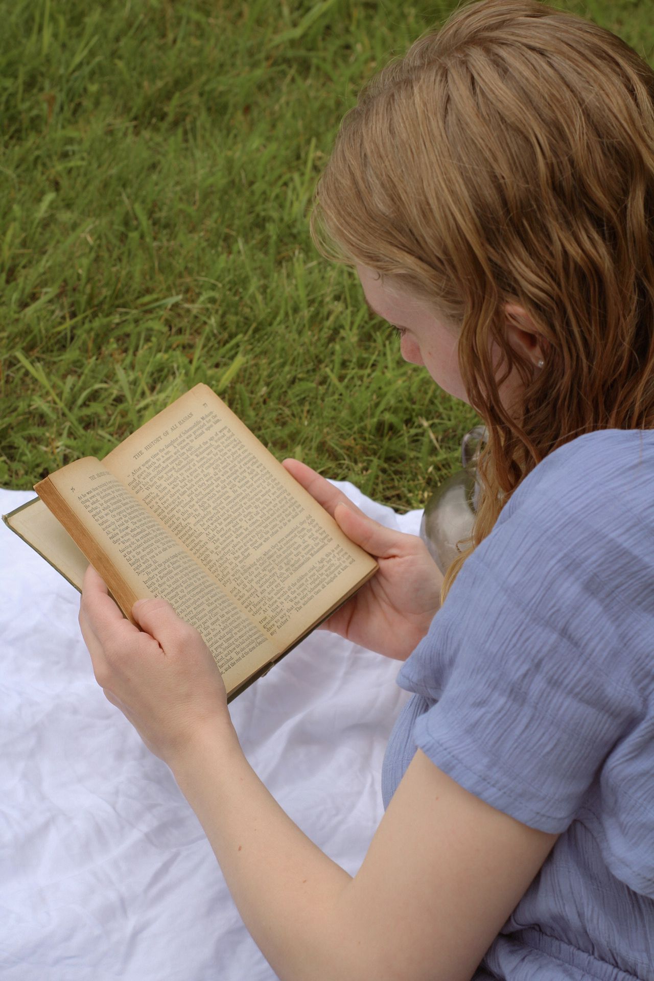 Woman in a blue dress enjoys a picnic on a white blanket with lanterns and old vintage books. Cottage core vibes